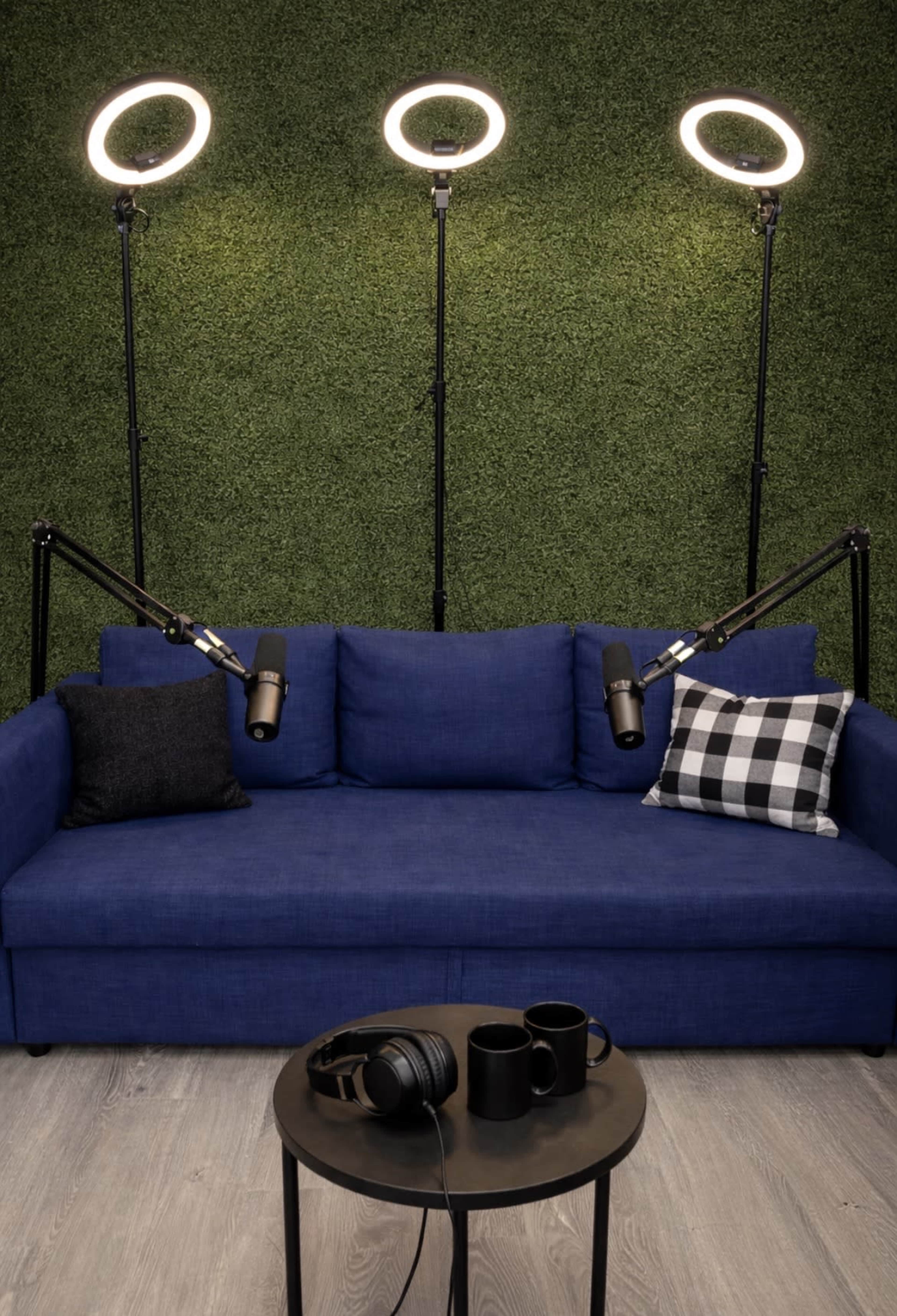 A blue couch with black and white patterned pillows is positioned in front of three illuminated ring lights, with a small table featuring headphones and two mugs nearby.