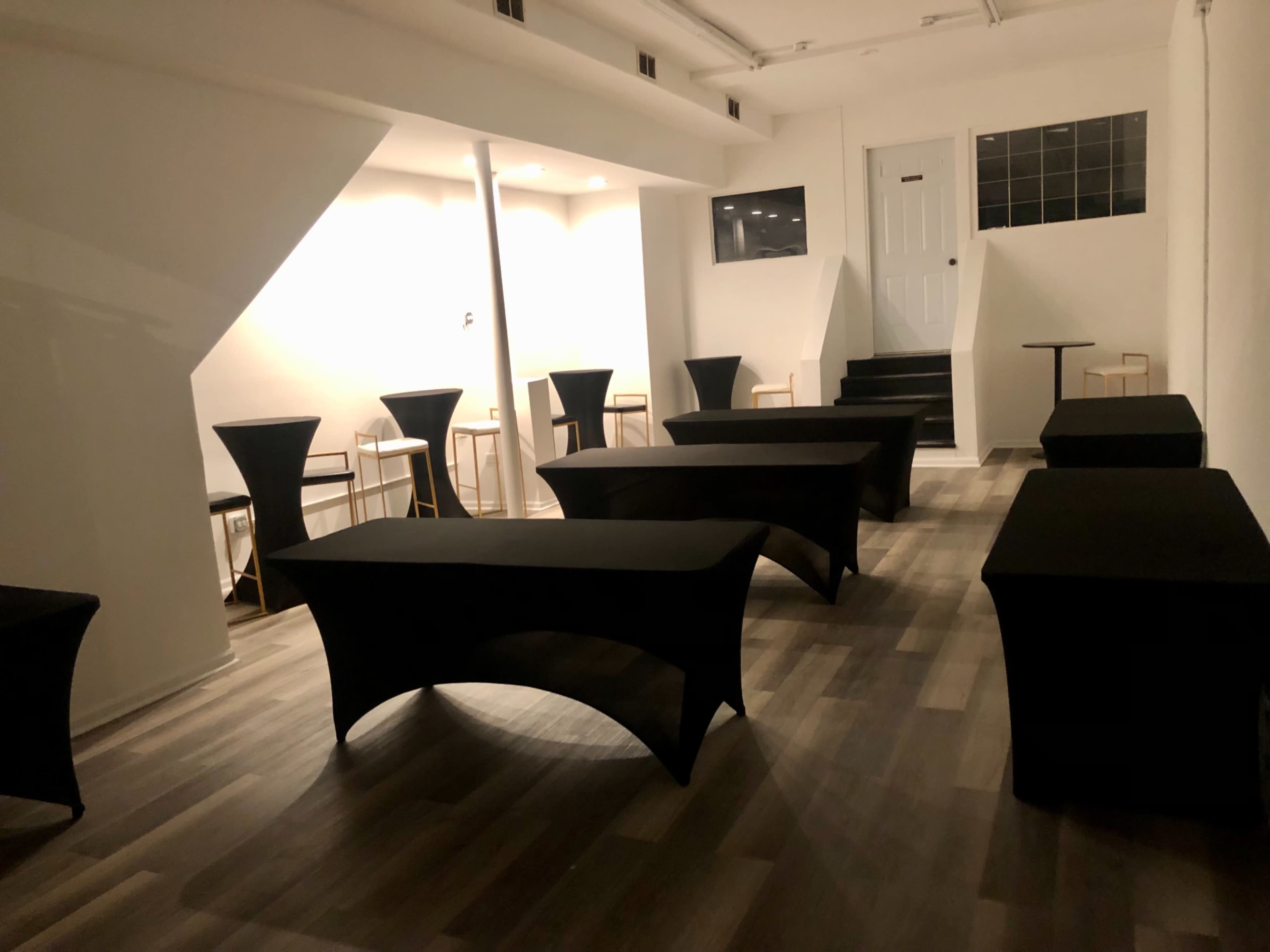The image shows a sparsely furnished room with several black-covered tables and a staircase leading to a door at the back.