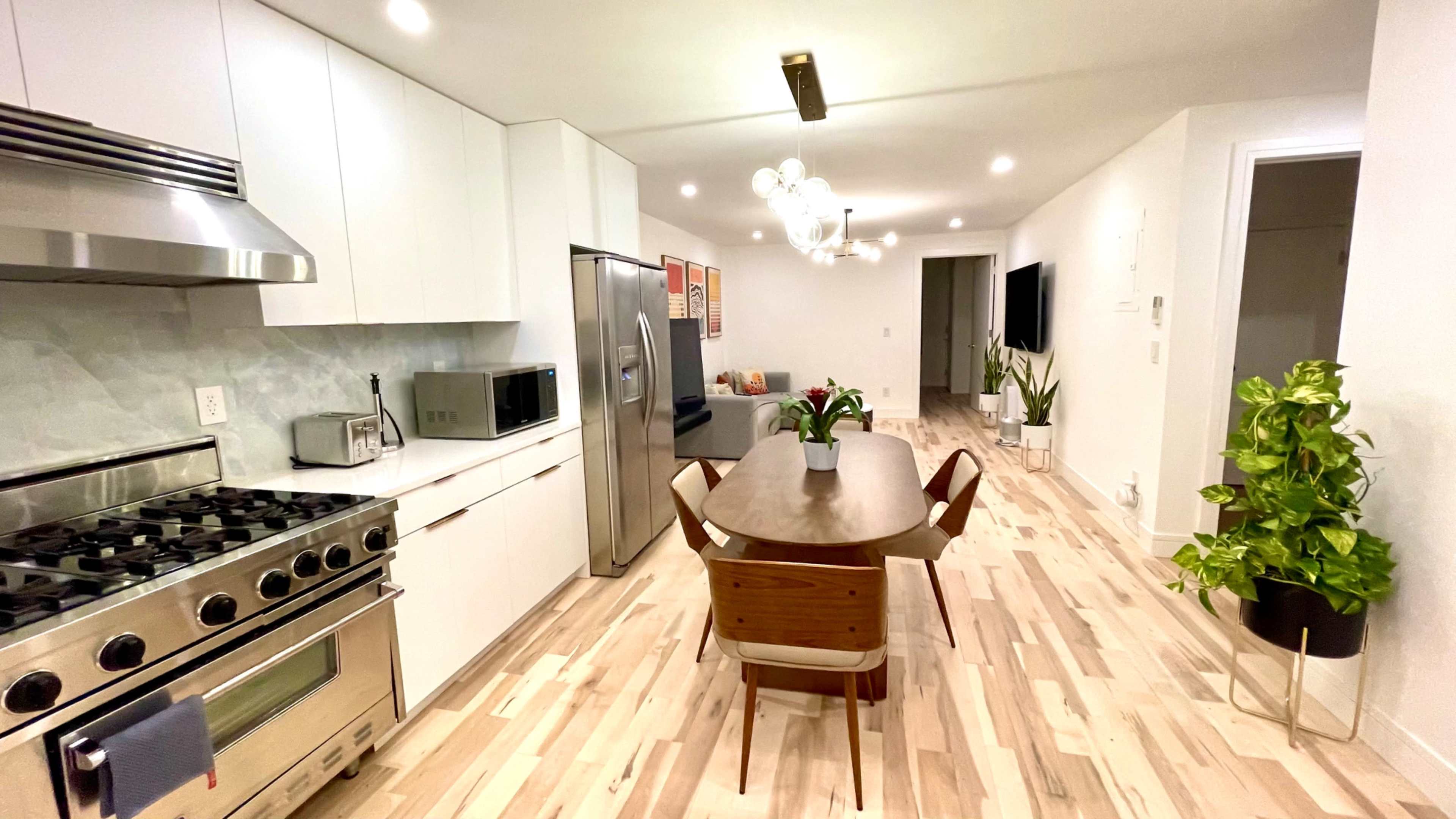 The image shows a modern kitchen and dining area with sleek white cabinetry, stainless steel appliances, a wooden dining table, and contemporary light fixtures.