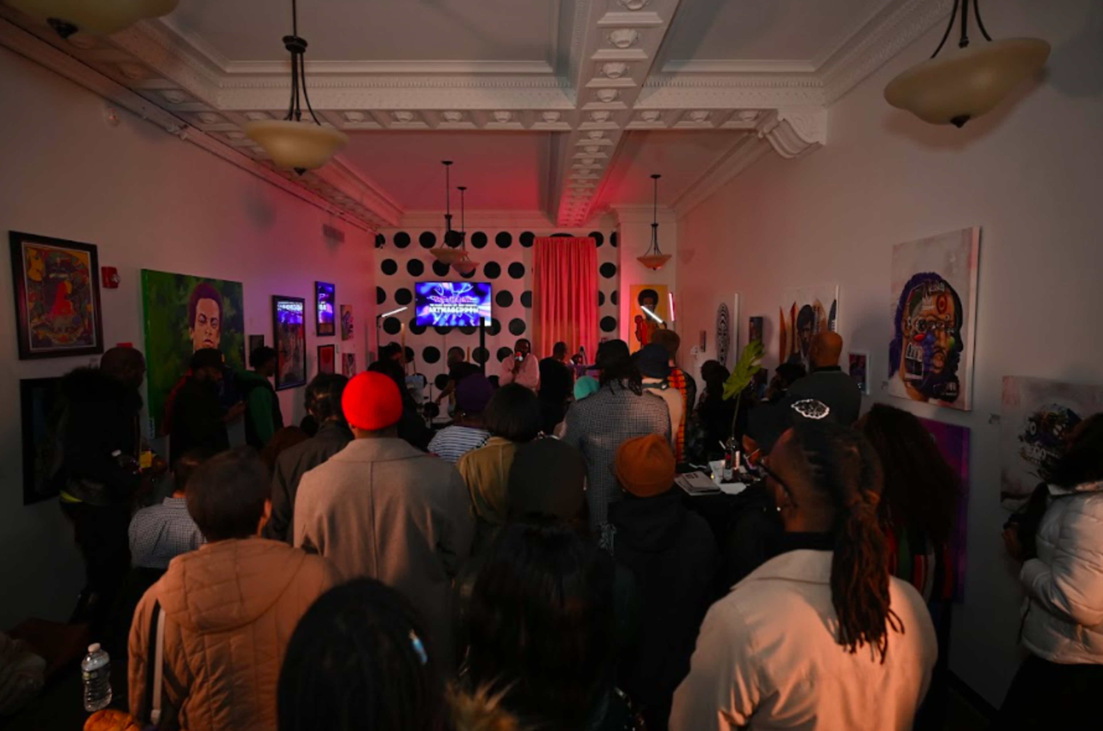 A crowd of people gathers in a dimly lit room adorned with colorful artwork, facing a screen for a performance or presentation.