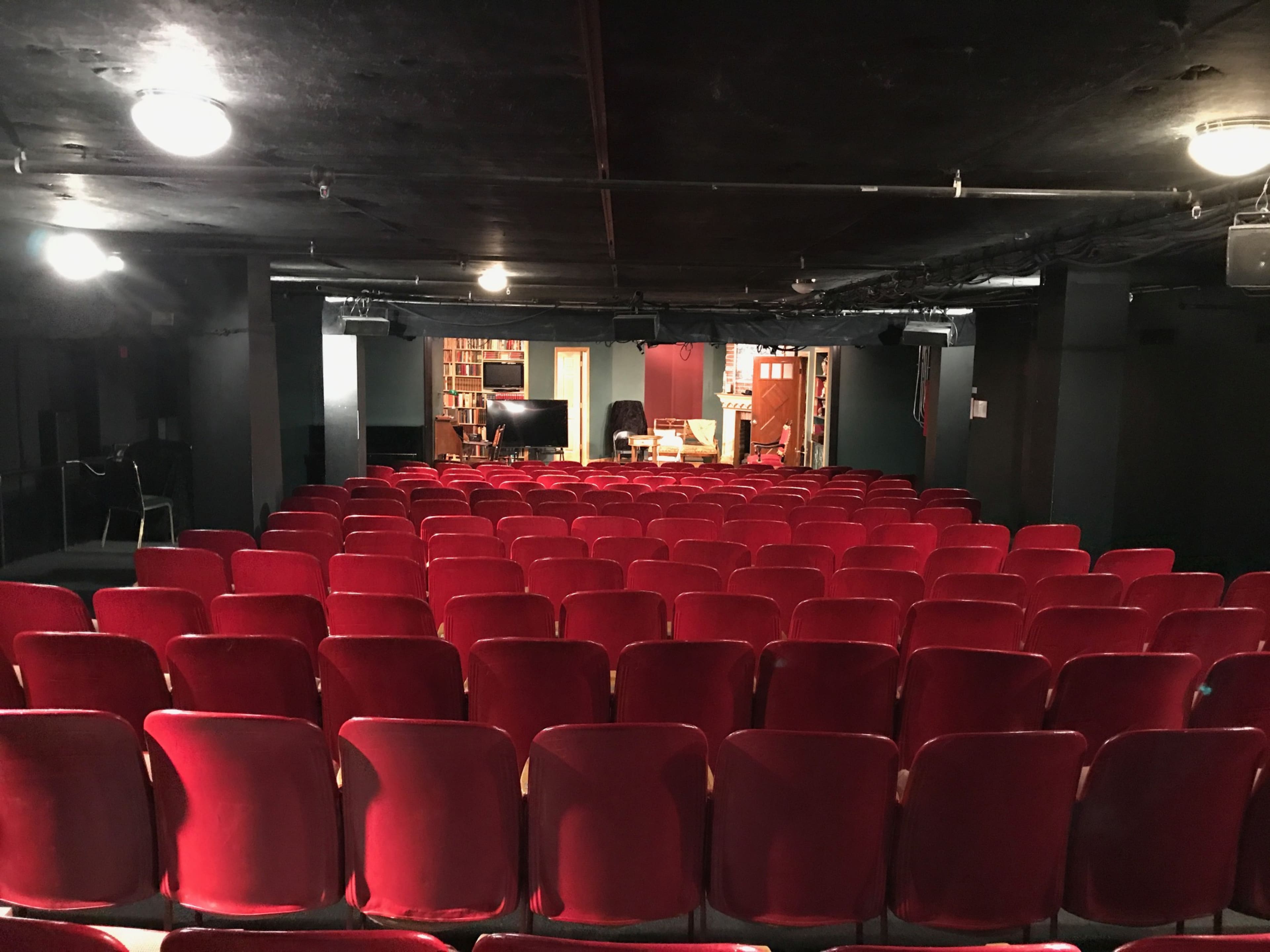 The image shows an empty, dimly lit theater with rows of red velvet seats facing a stage set with shelves and furnishings in the background.