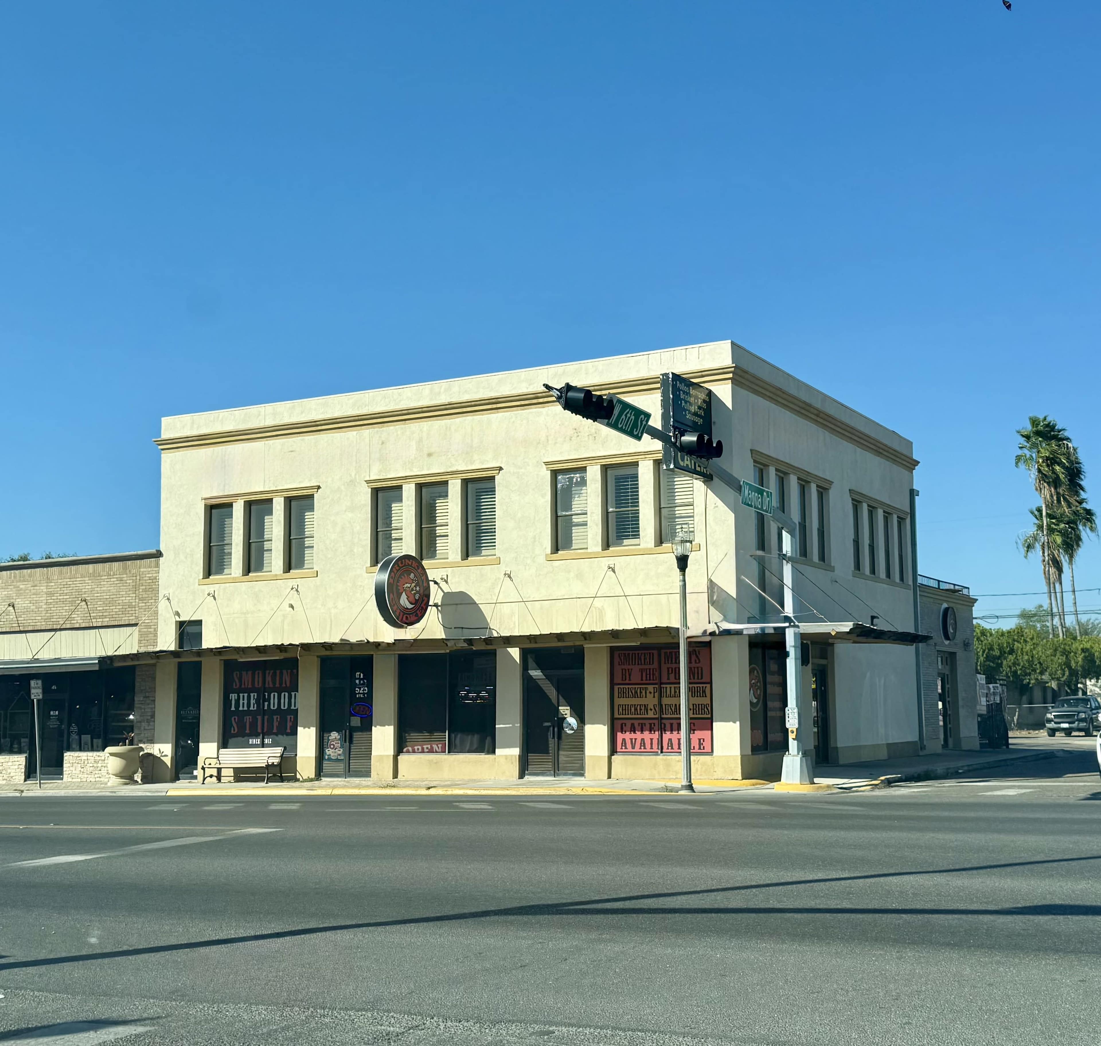 The image shows a two-story commercial building at a street corner, with various storefronts visible on the ground level and palm trees in the background.