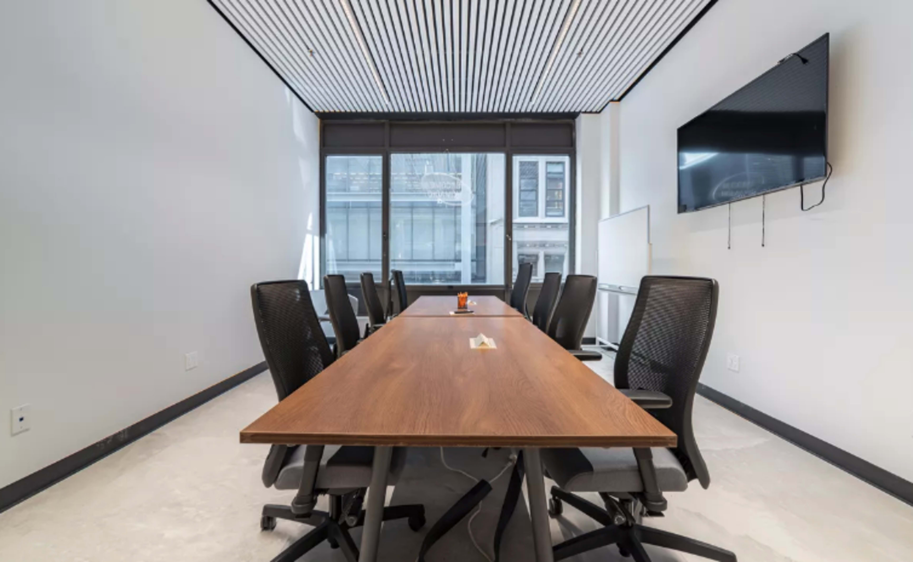 A conference room features a long wooden table surrounded by black mesh chairs and a wall-mounted television, with large windows allowing natural light.