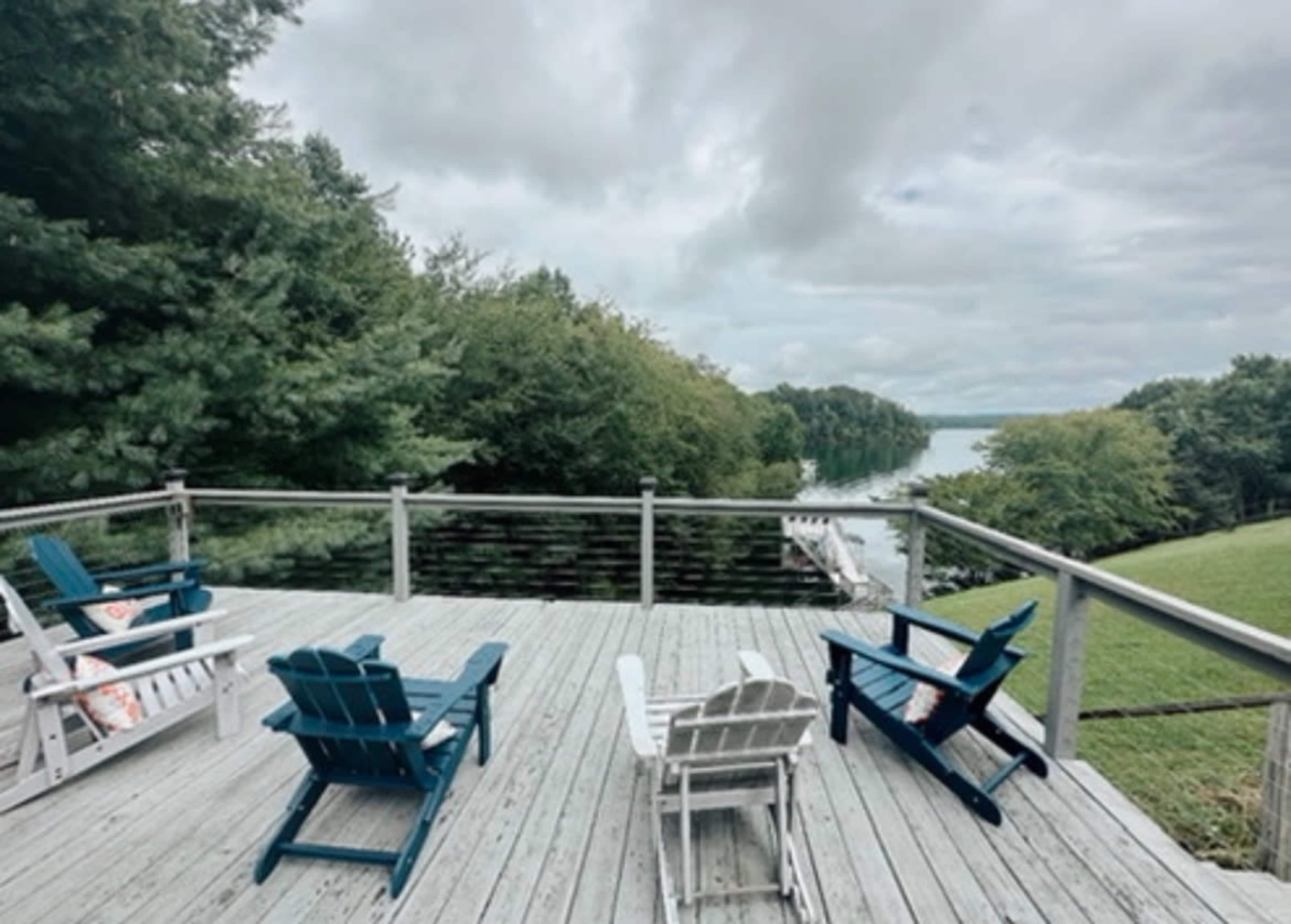 A wooden deck with four colorful Adirondack chairs overlooks a calm river surrounded by greenery.