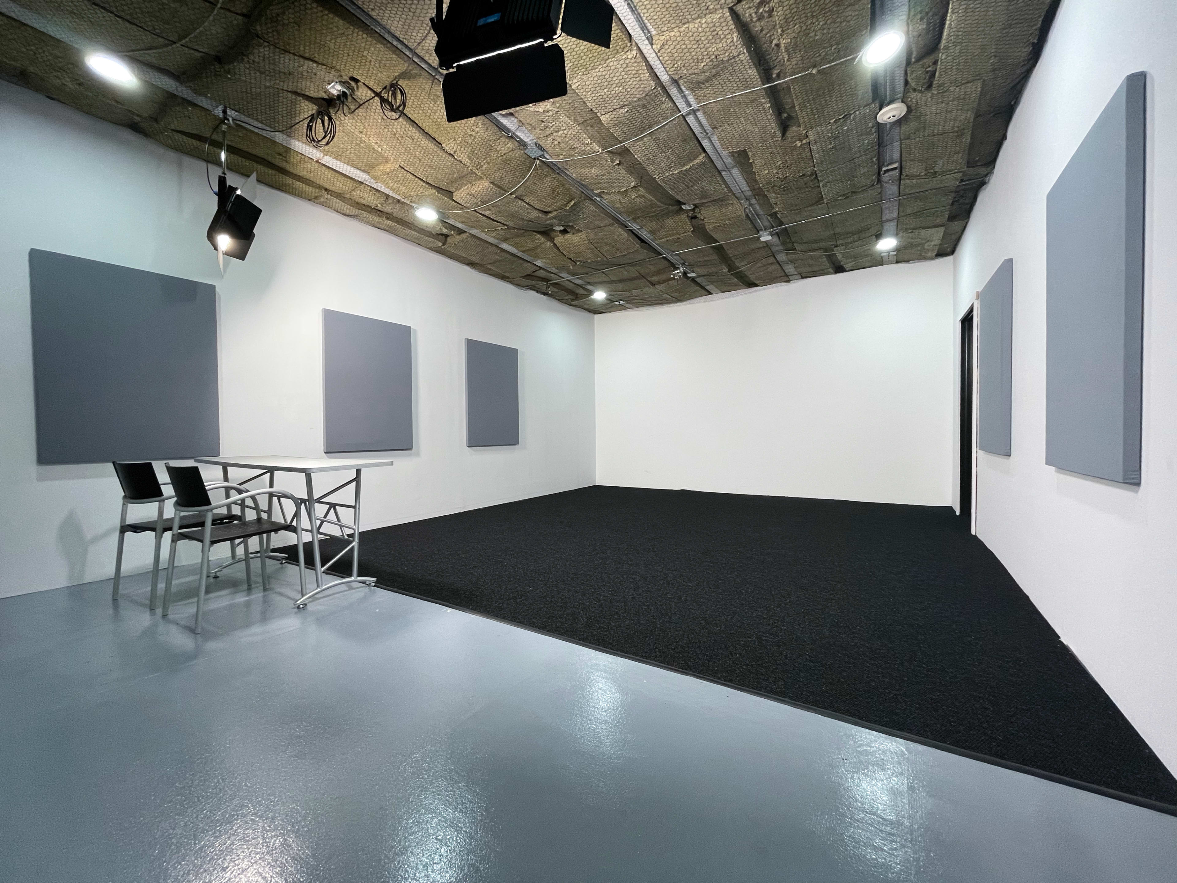 The image shows an empty room with a dark carpeted floor, a table with two chairs, and soundproofing panels mounted on the white walls.
