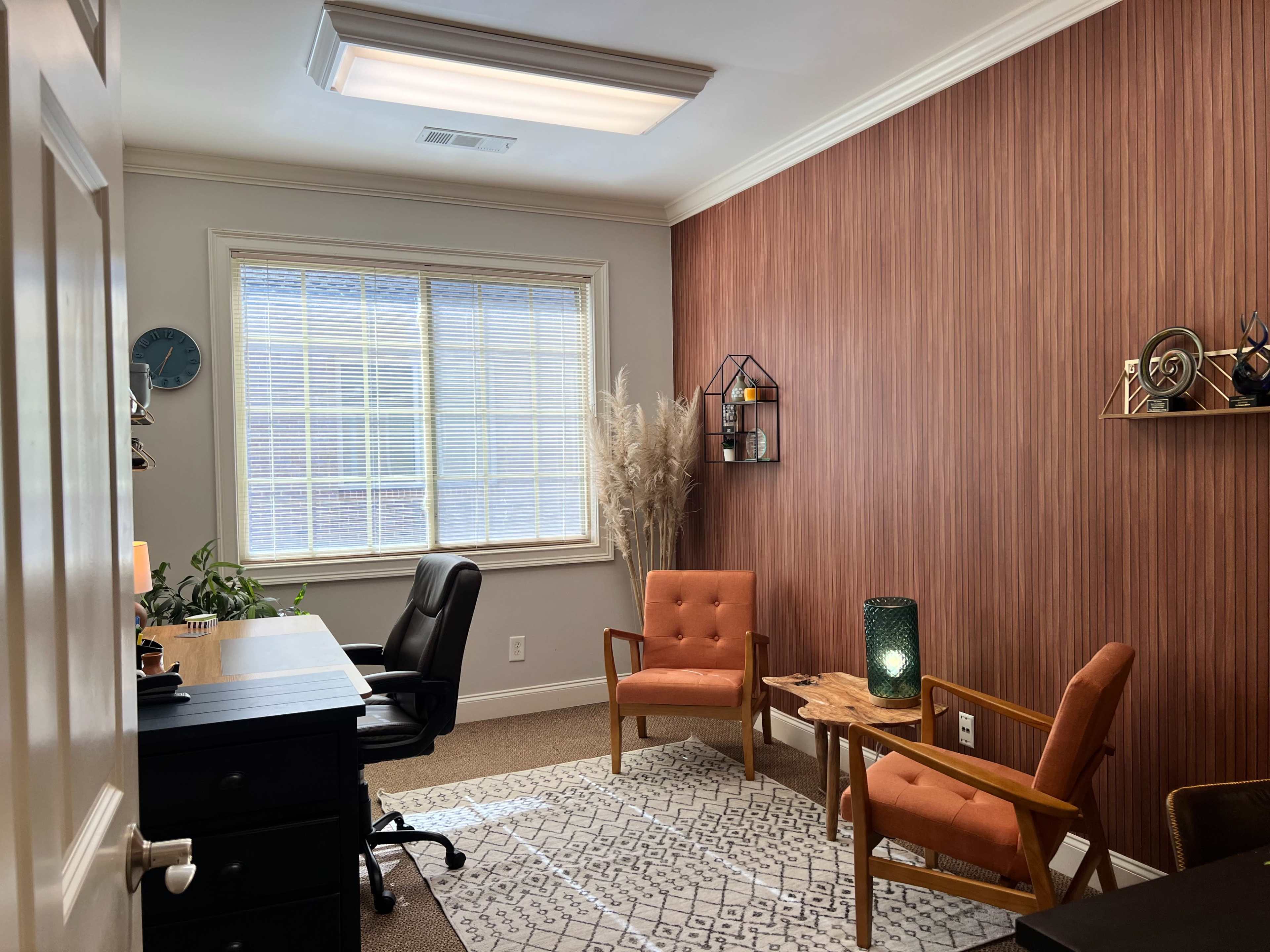 The image shows a well-lit office space featuring two orange chairs, a desk with a black chair, and decorative elements including a plant and wall hangings.