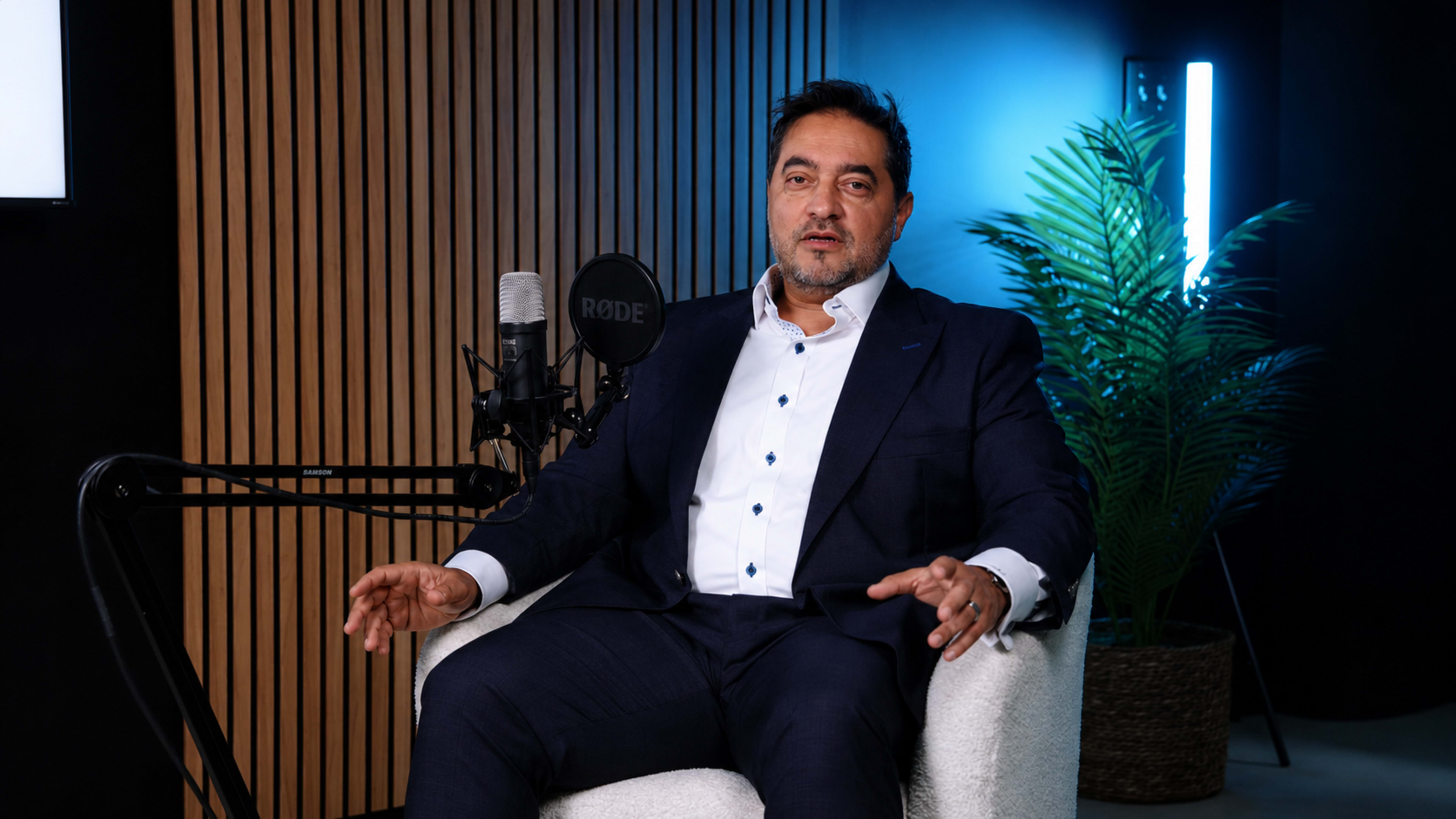 A man in a dark suit sits in a white armchair, speaking into a microphone in a modern studio setting with a plant and soft lighting.