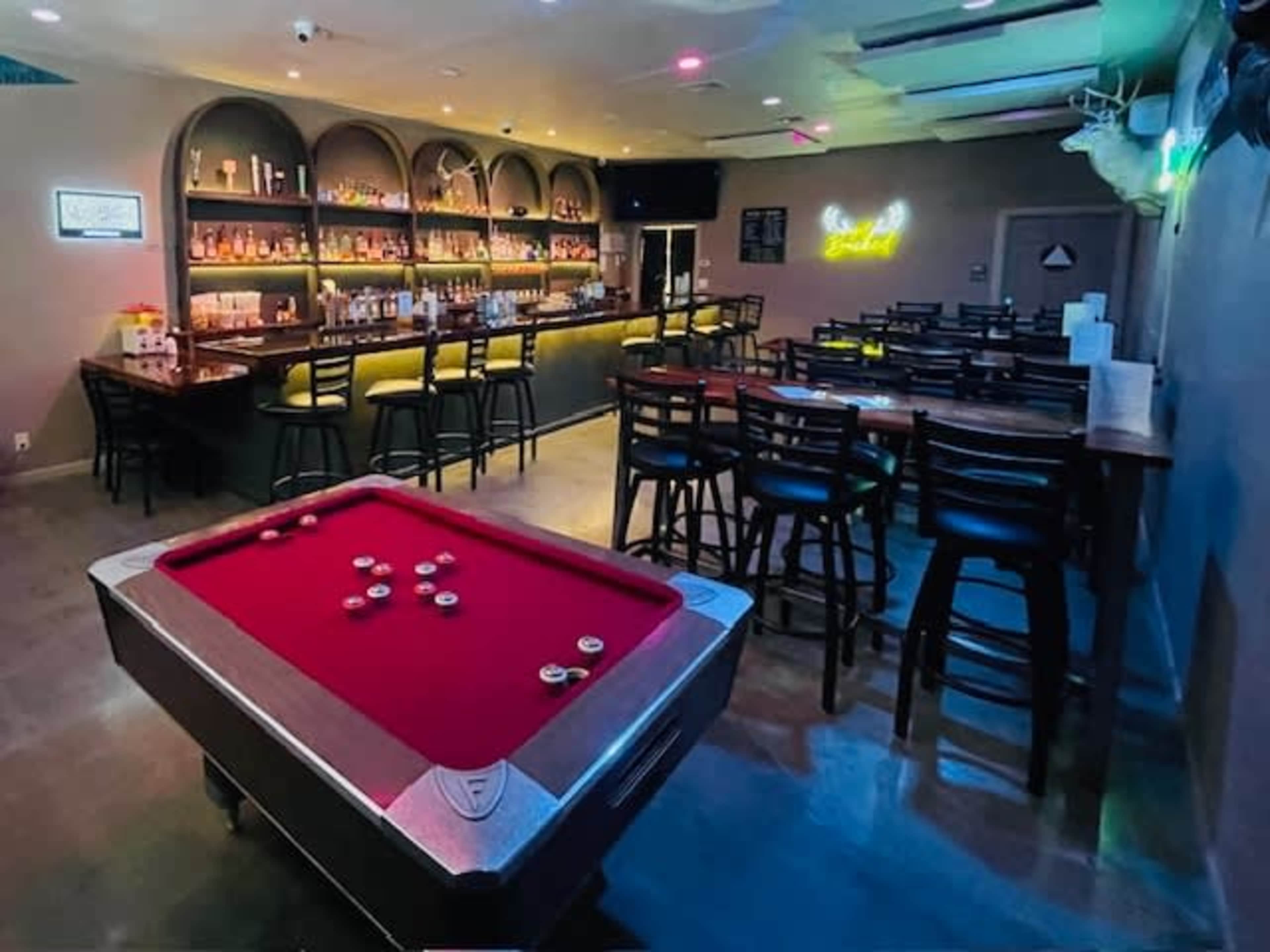 The image shows a bar area featuring a pool table in the foreground and shelves stocked with bottles in the background, illuminated by soft lighting.