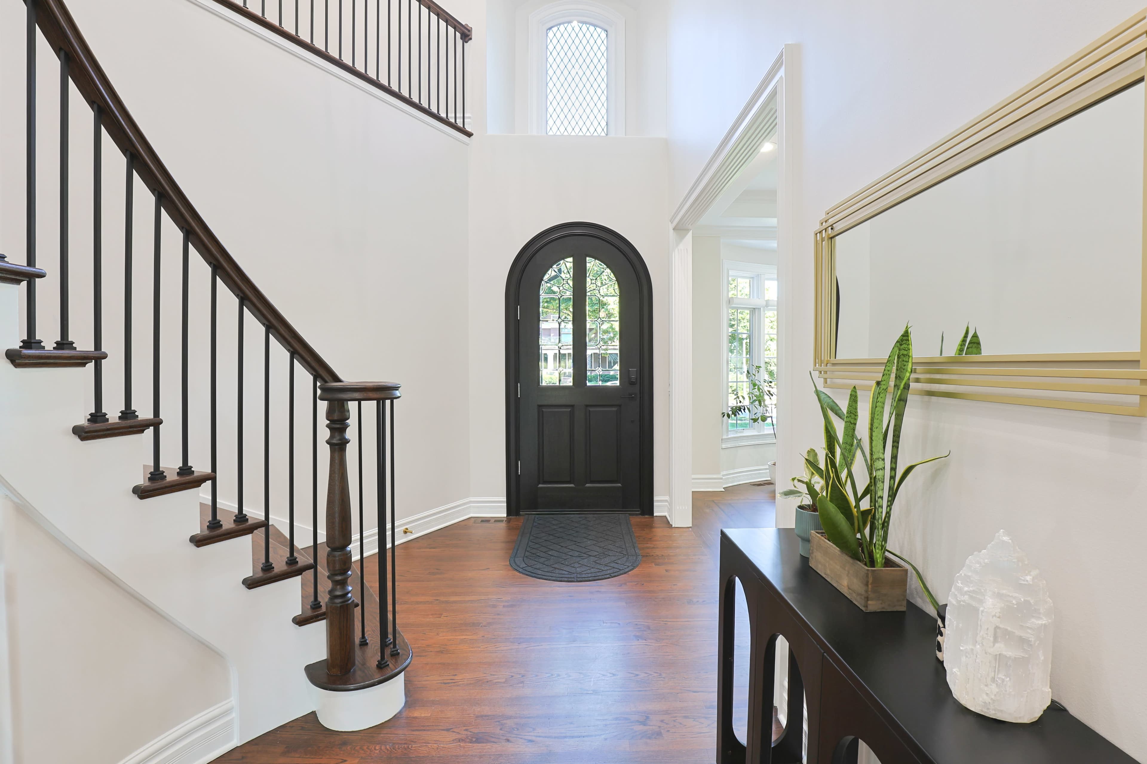 The image shows a spacious foyer with a black front door, a wooden staircase, and a console table adorned with plants and decorative objects.