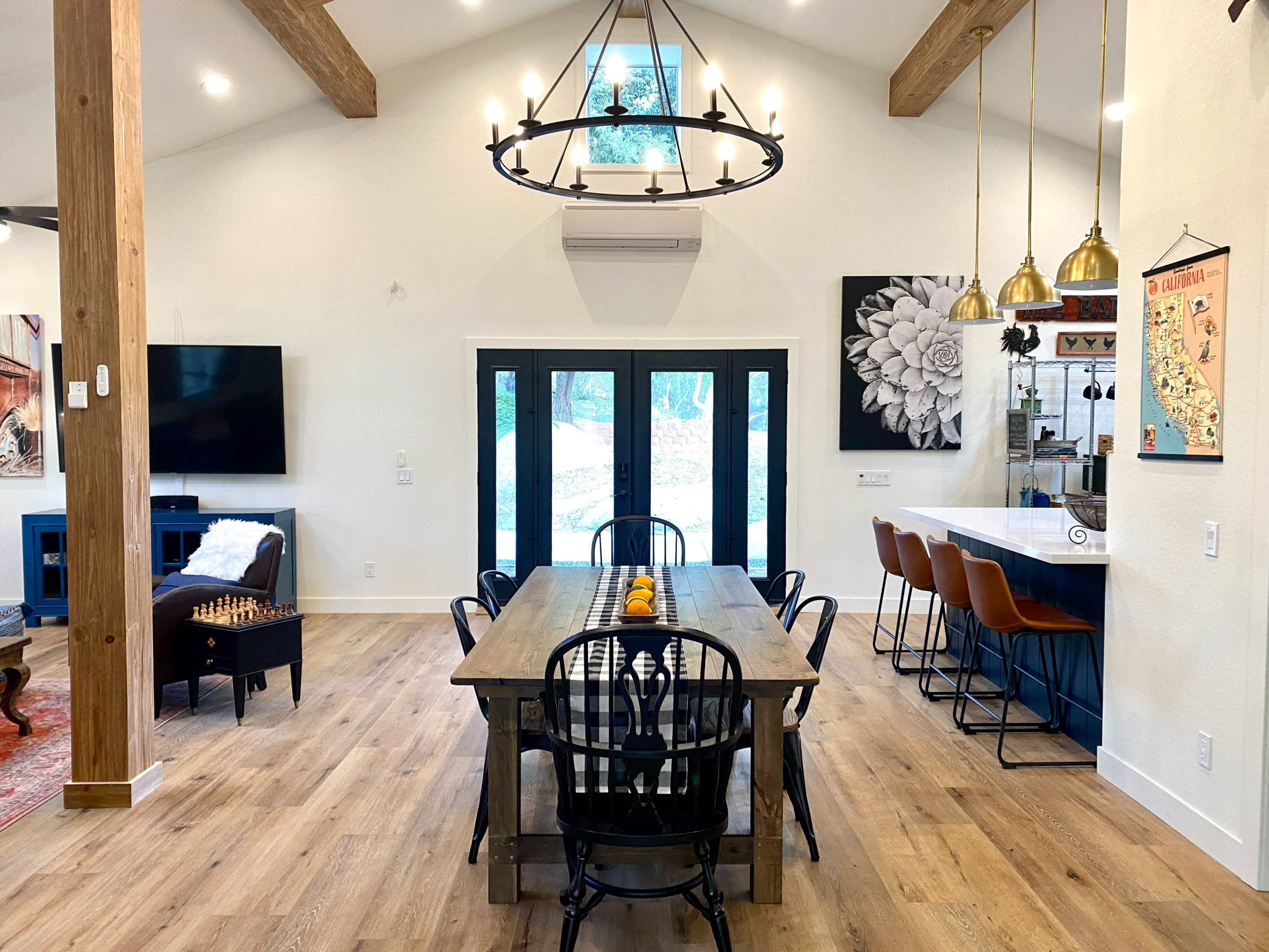 A spacious dining area features a long wooden table with black chairs, a chandelier overhead, and large windows overlooking an outdoor view.