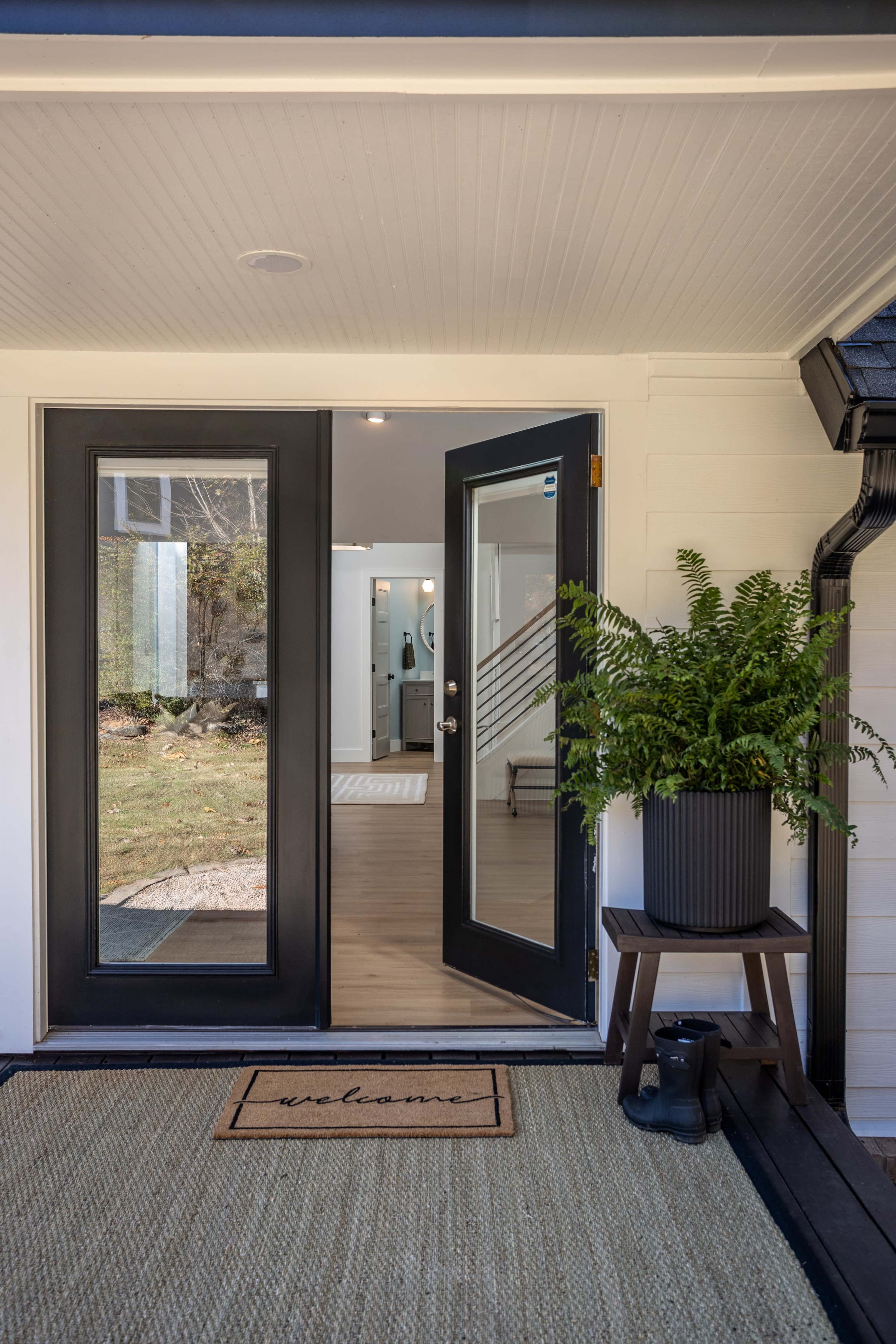 The image shows a modern front entrance with double doors open to a bright interior, featuring a welcome mat and a potted plant on a side table.