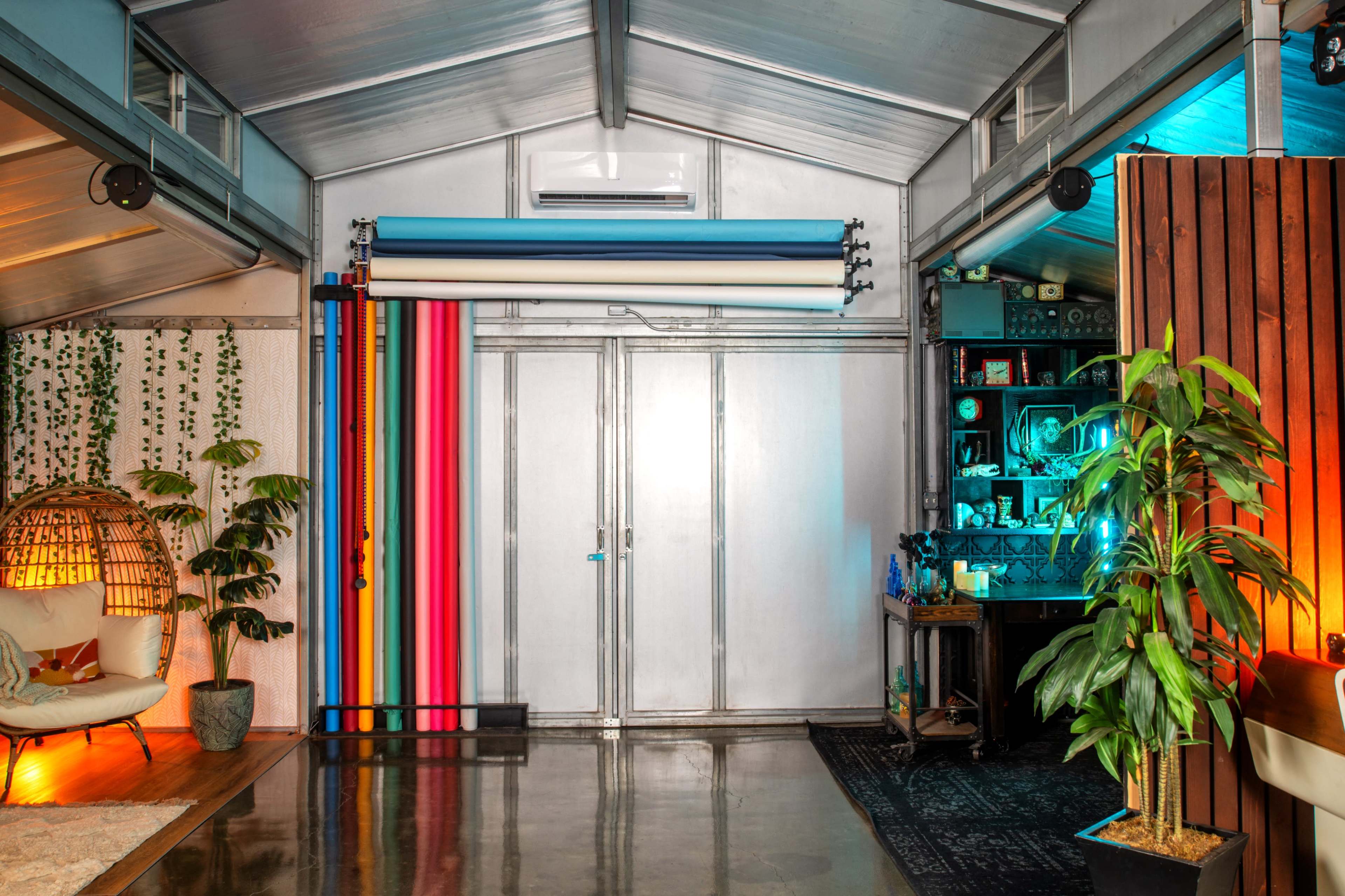 The interior of a modern studio features colorful backdrop rolls, plants, and a cozy seating area, with lighting illuminating one side of the room.
