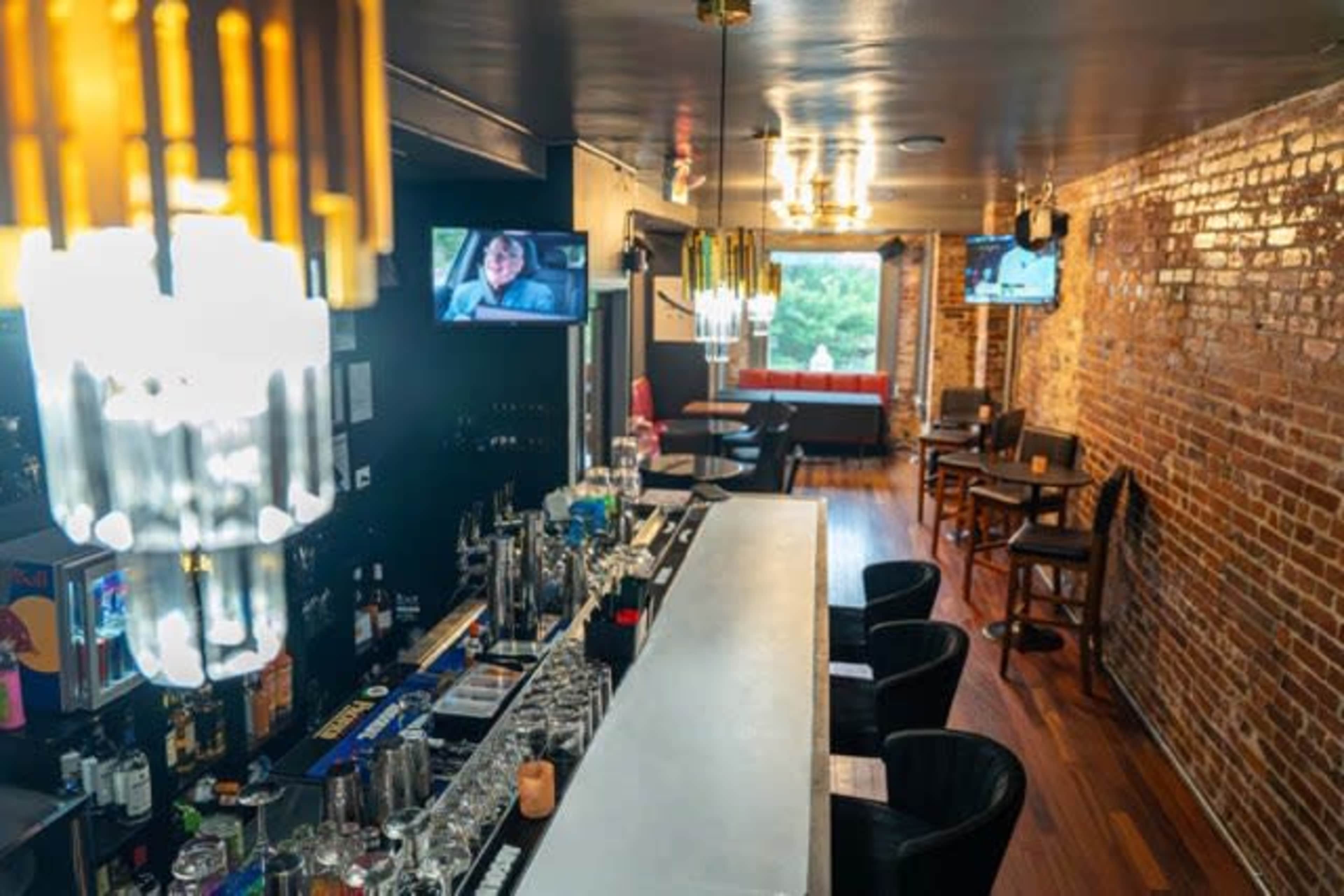 A modern bar scene features a long white countertop with various drinks positioned on it, surrounded by black chairs, and two televisions displaying content on the walls of a brick interior.
