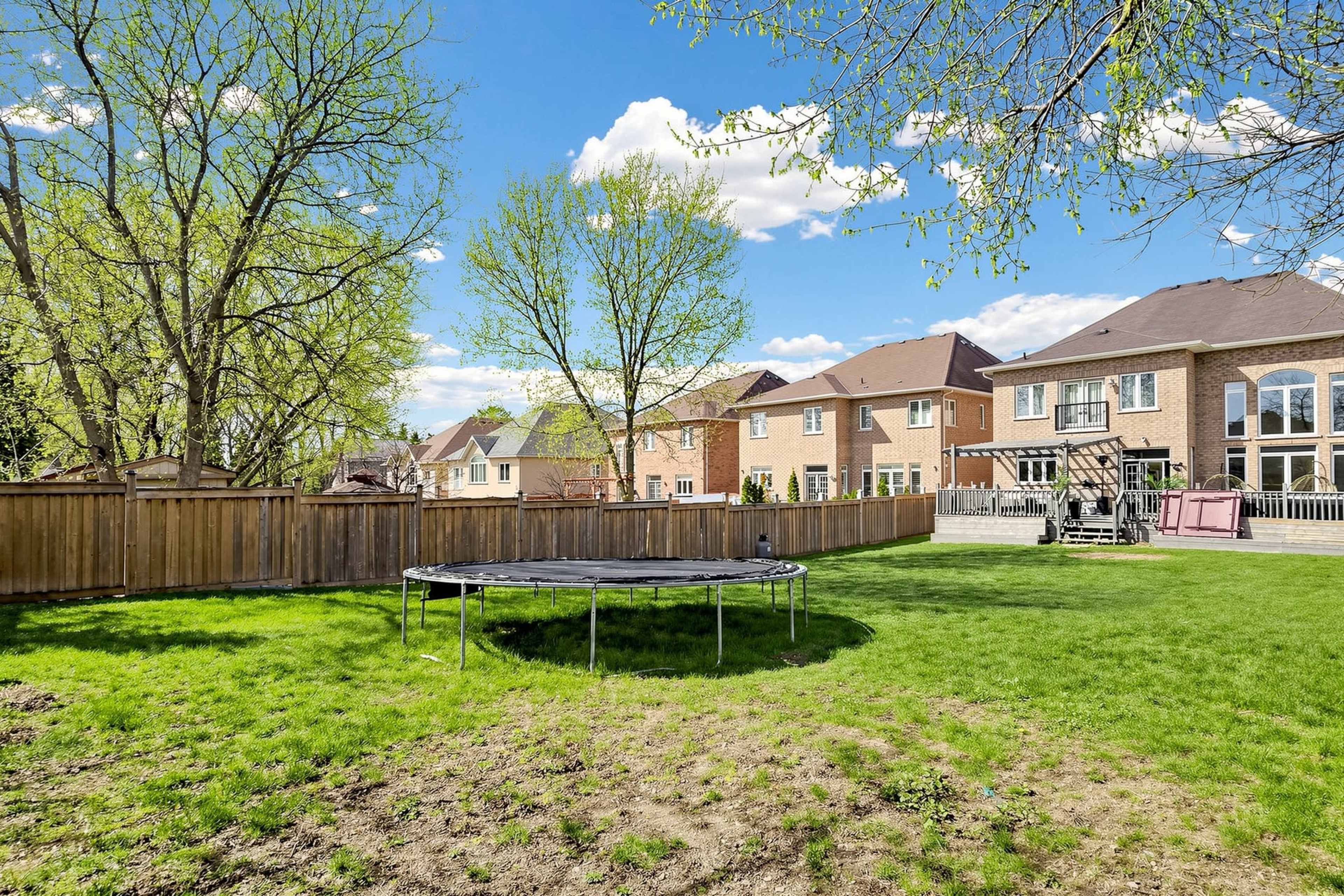 A spacious backyard features a trampoline surrounded by lush green grass and several houses with wooden fences in the background.