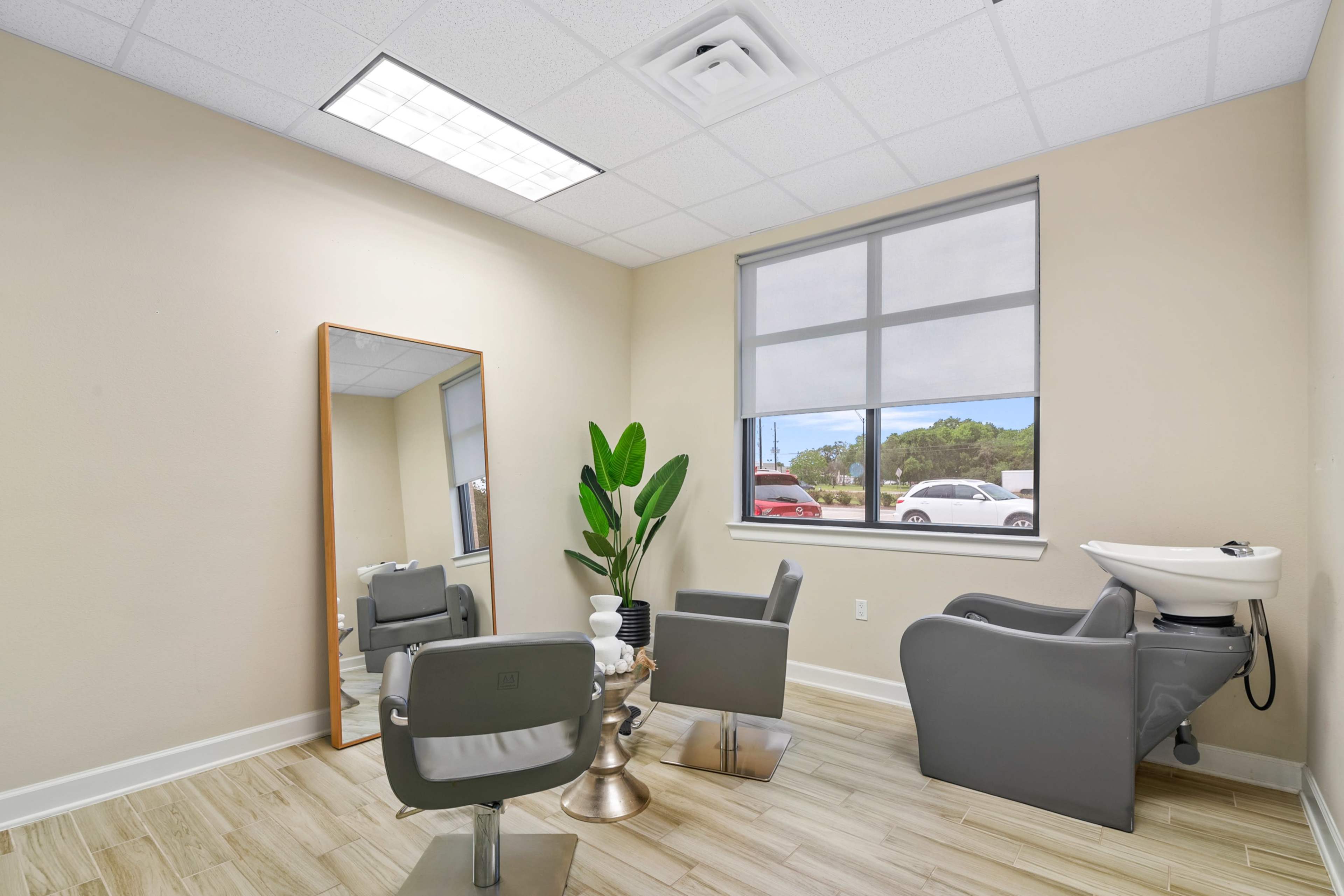 A modern salon interior with two gray chairs, a large mirror, a potted plant, and a hair wash station beside a window.