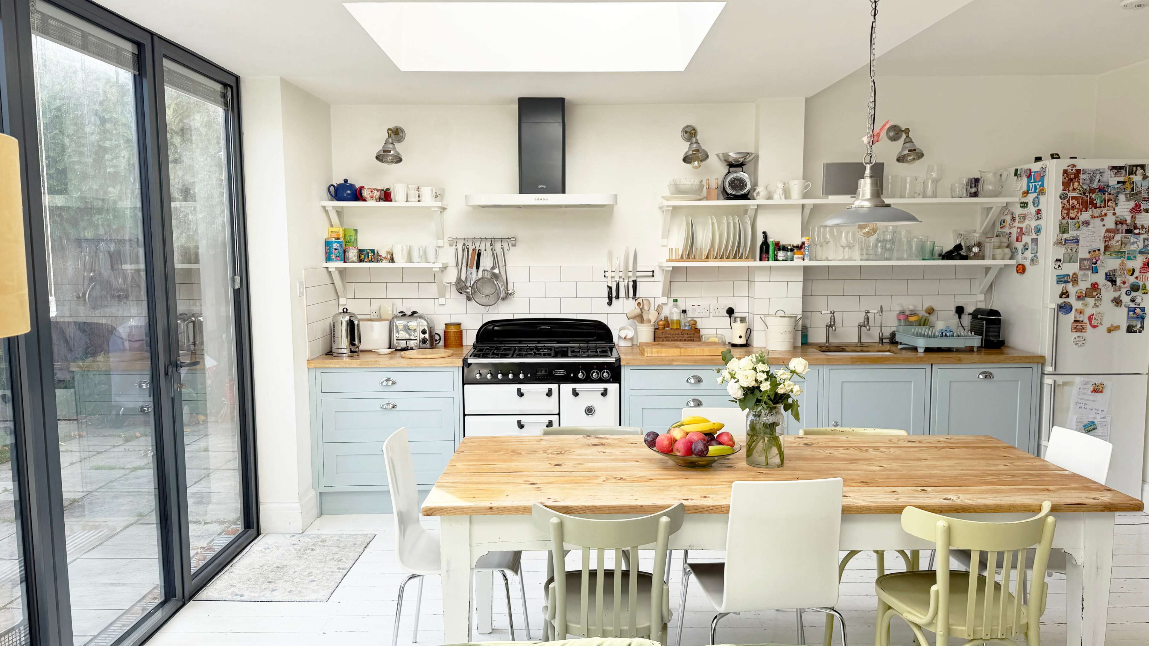 A modern kitchen features a central wooden table surrounded by colorful chairs, with a black stove and shelves filled with kitchenware against a white-tiled wall.