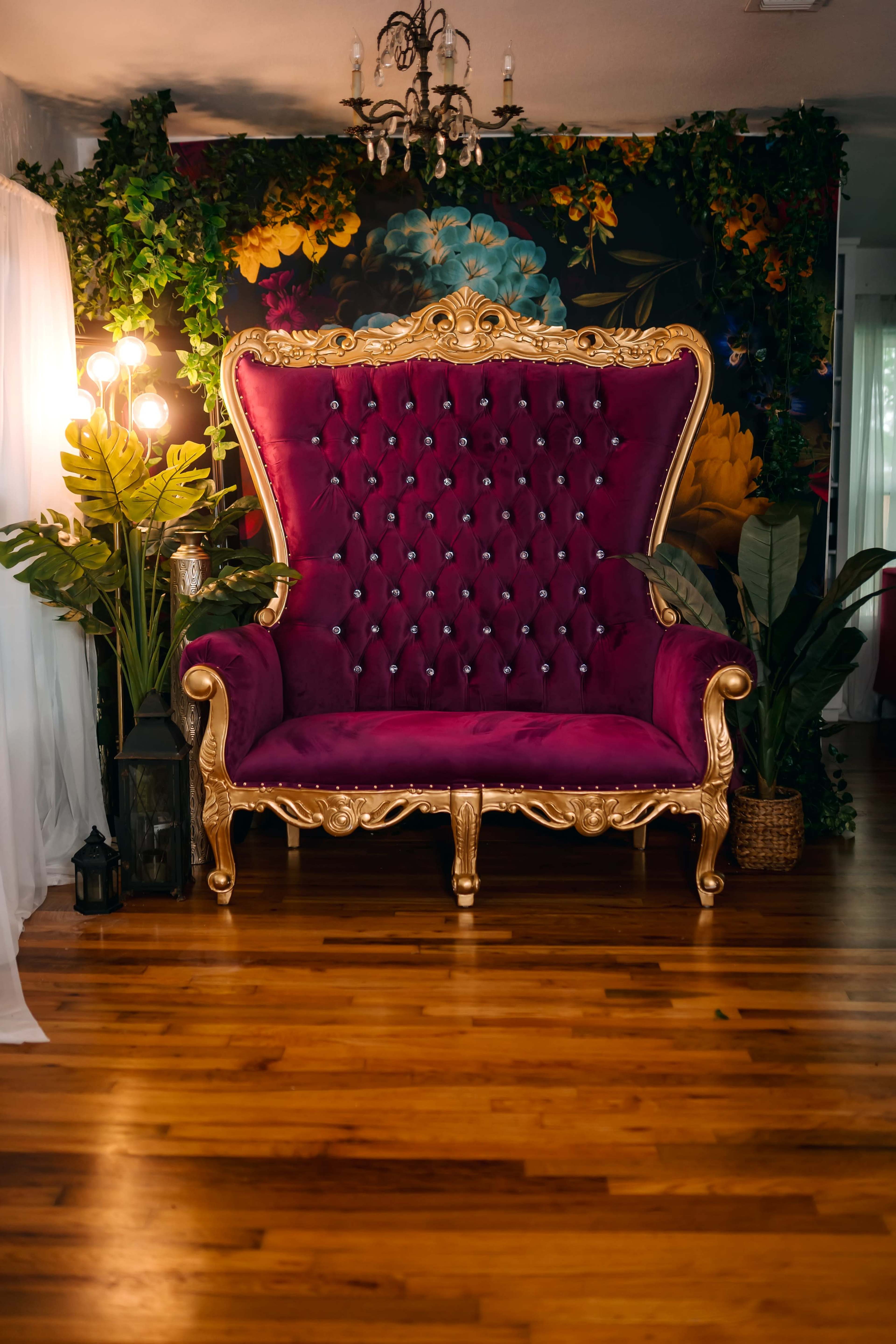 The image features a large purple velvet throne with gold accents, set against a vibrant floral backdrop and accompanied by decorative plants and ambient lighting.