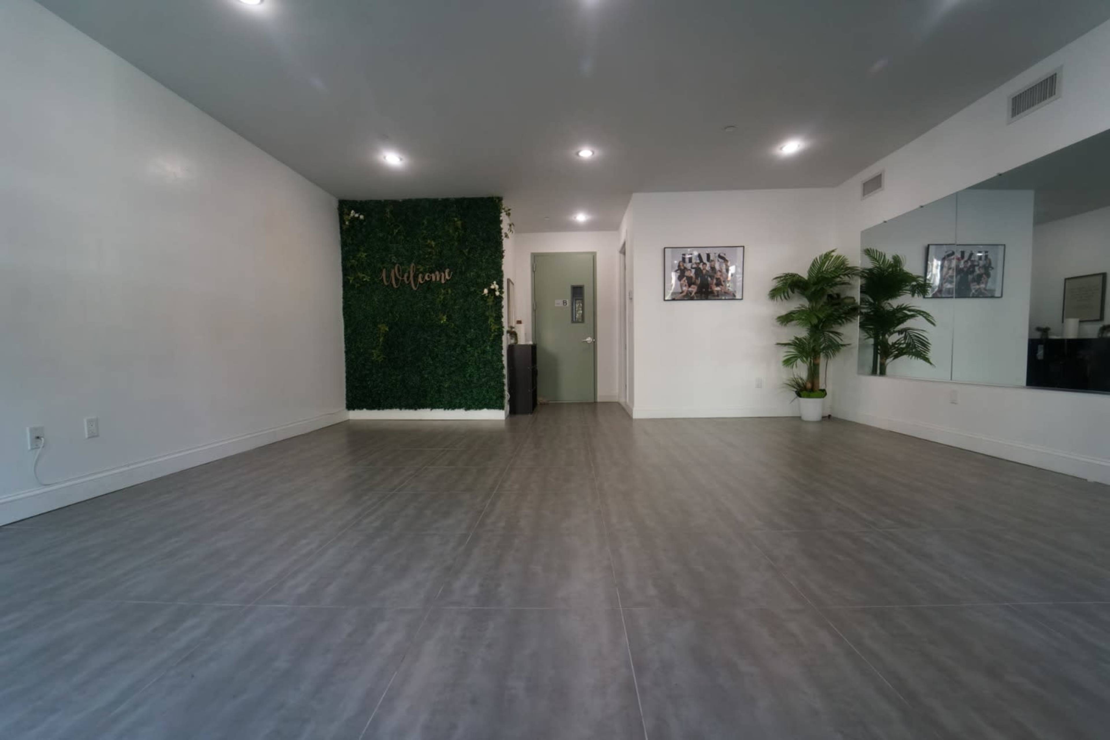 An empty room with grey tile flooring, a welcome-themed green wall, a mirror, and a door at the far end.