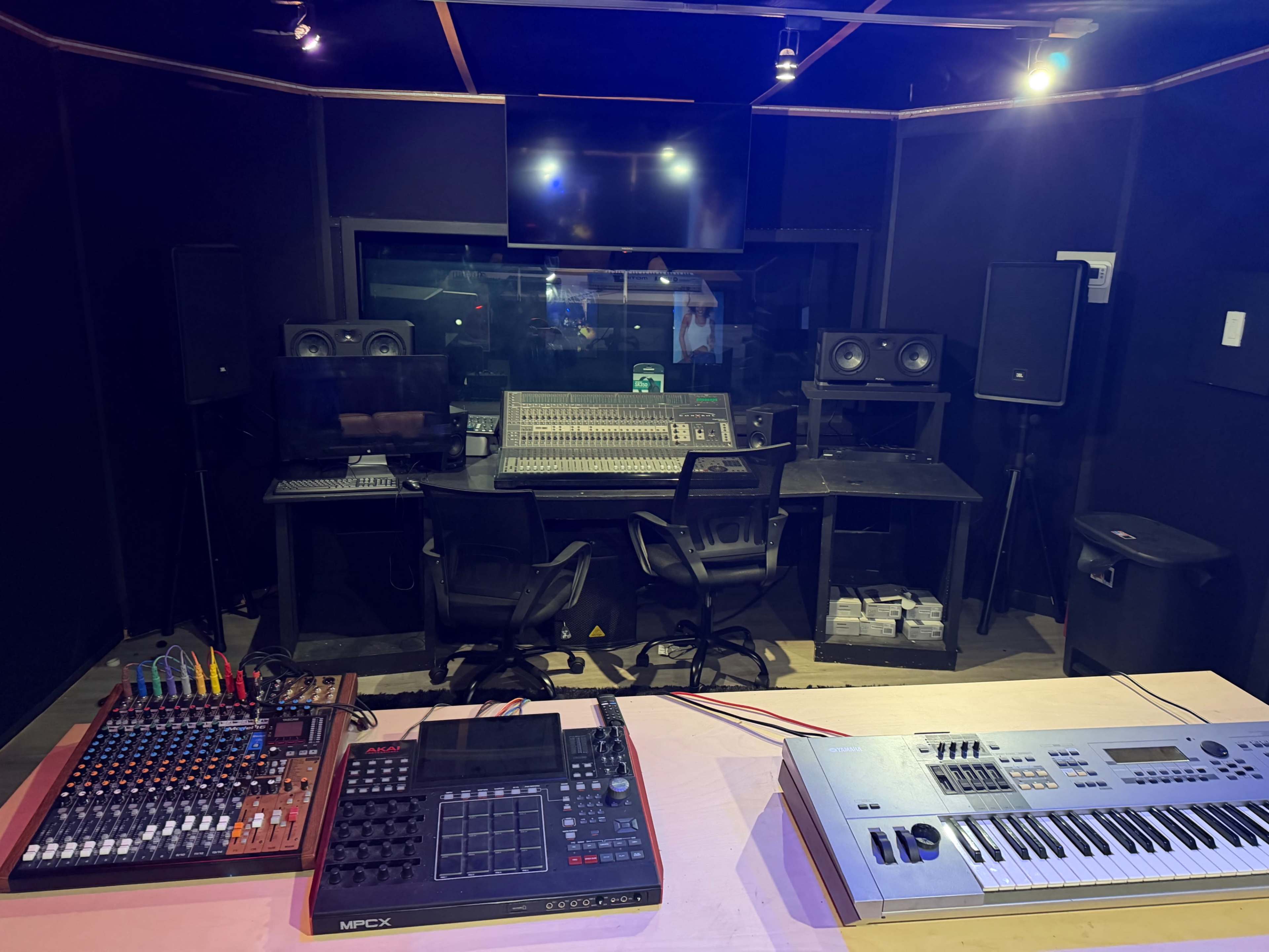 The image shows a recording studio with a mixing console, speakers, a drum machine, and a keyboard, all set up in a darkened environment.