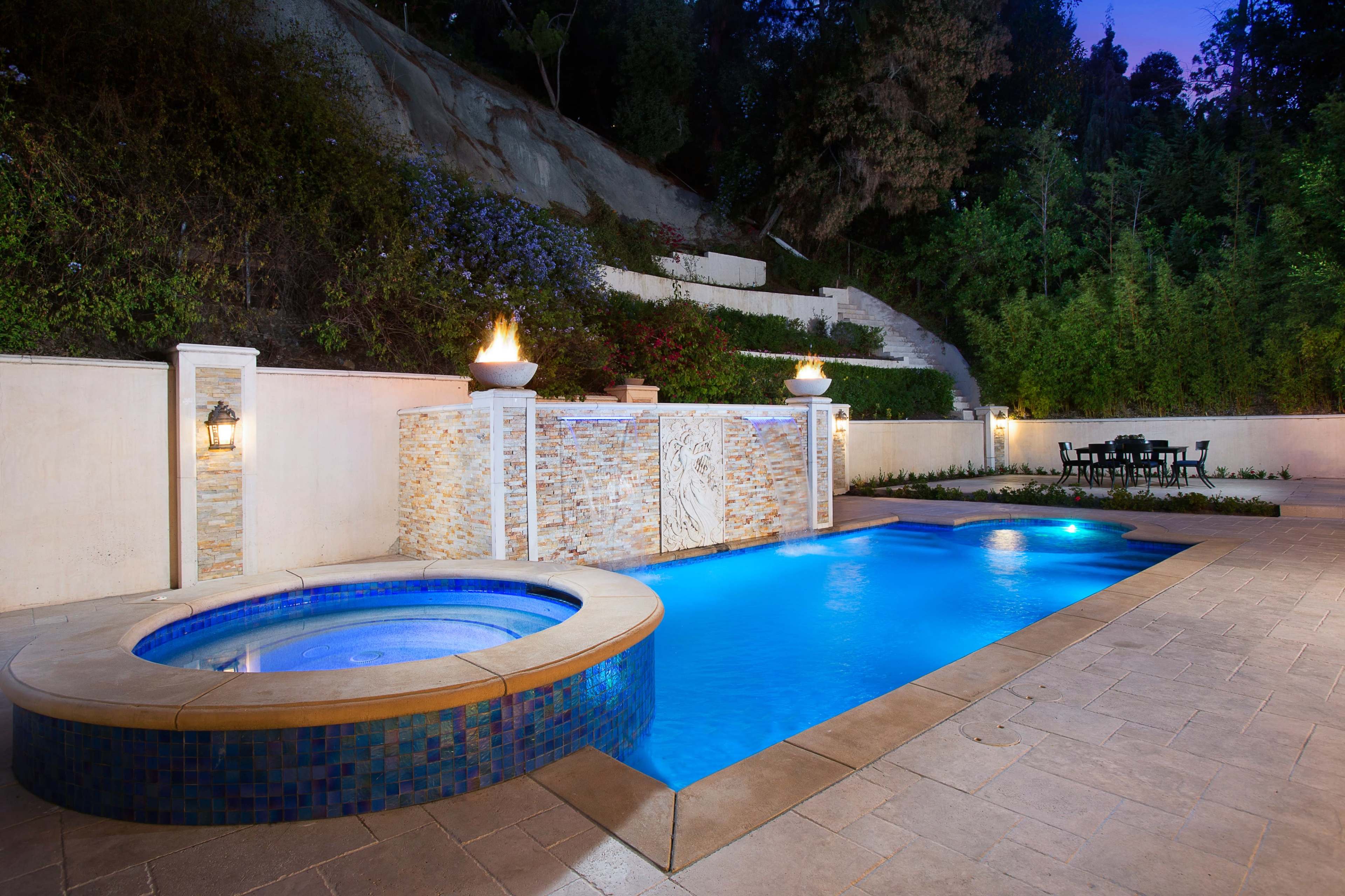 The image shows a backyard space featuring a pool and a spa, with stone wall accents and seating arranged nearby in a landscaped area.