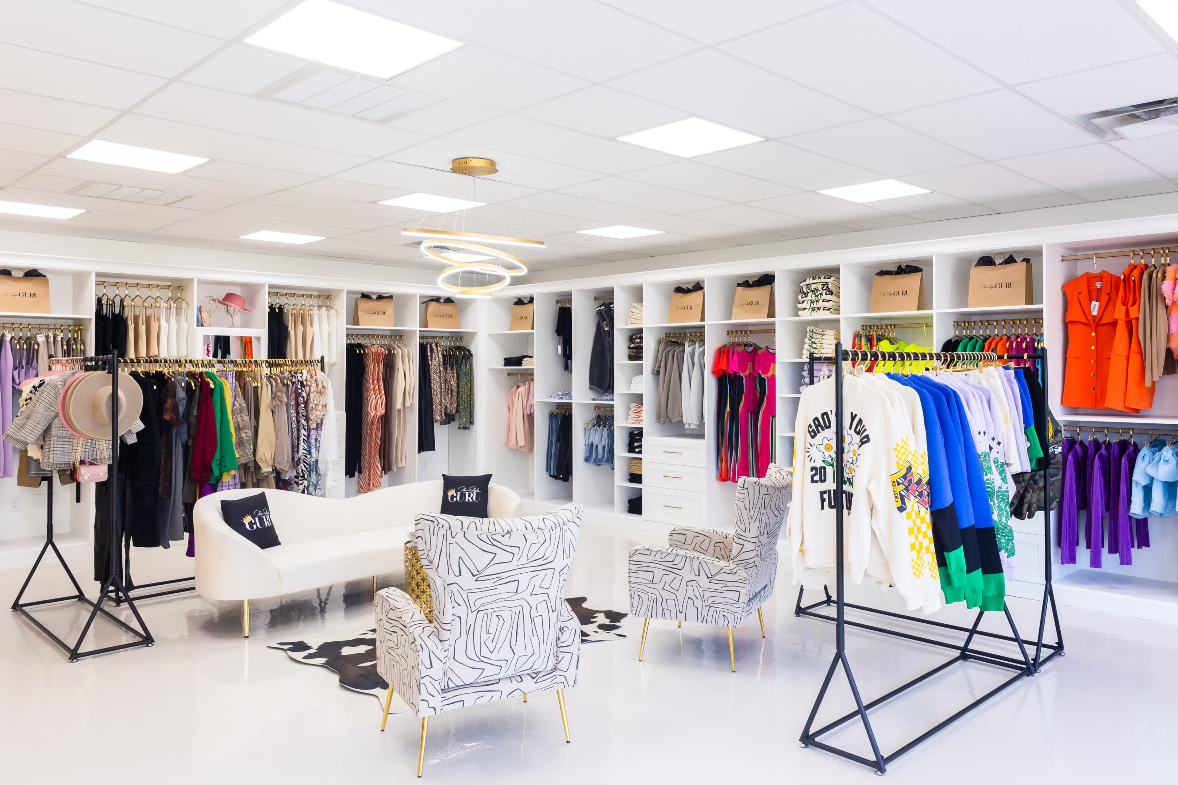 A brightly lit clothing boutique featuring organized racks of apparel, a seating area with two patterned chairs, and displays of accessories.