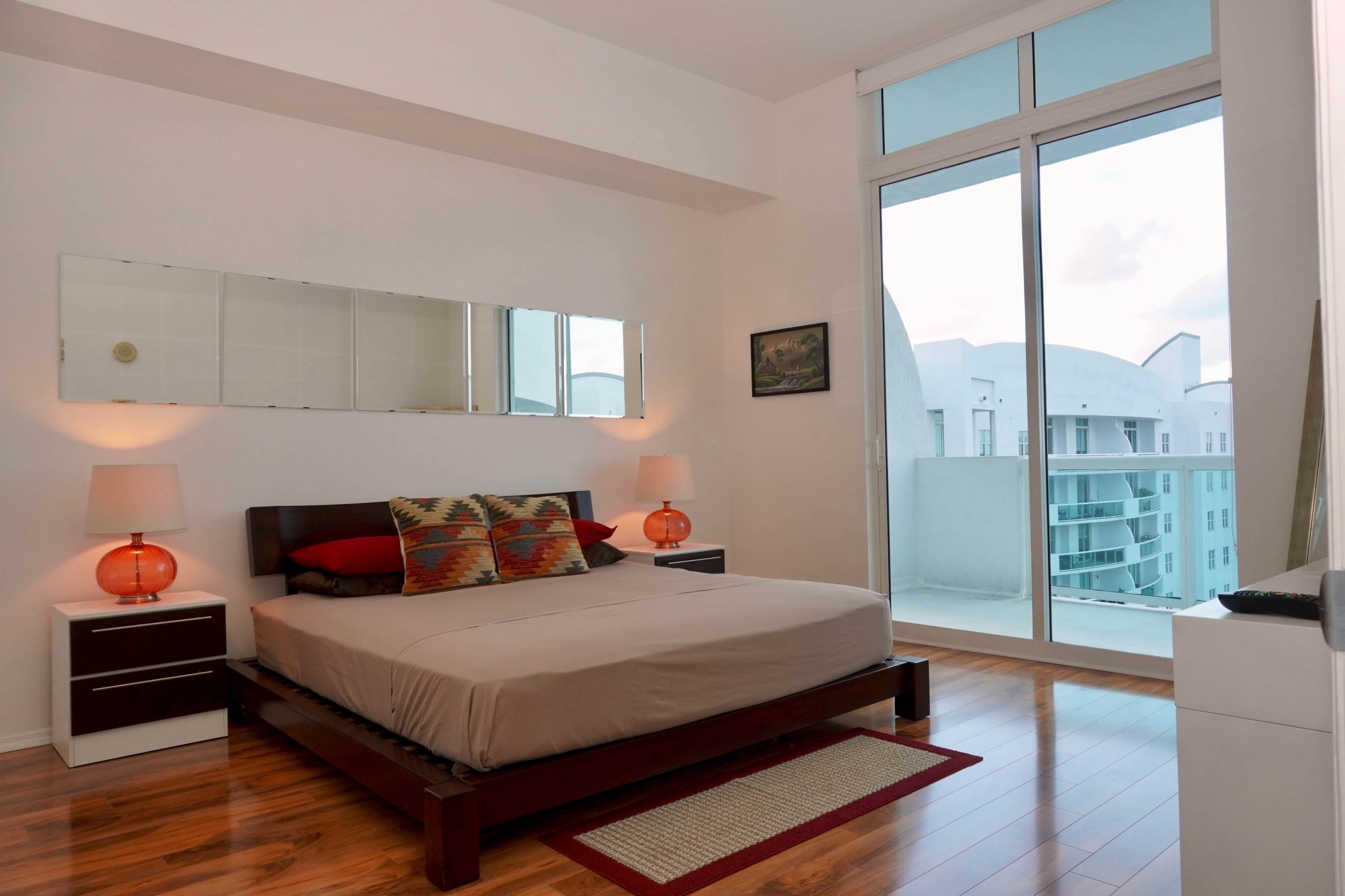 A modern bedroom features a bed with decorative pillows, two nightstands with lamps, and a large window overlooking a balcony.