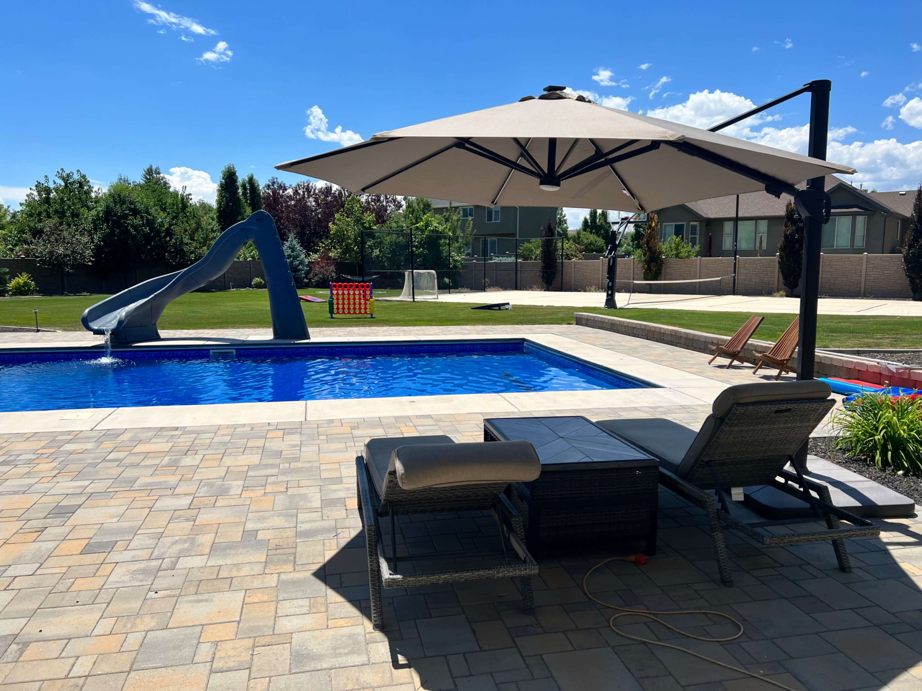 A backyard pool area featuring a blue water slide, a shaded seating area, and lounge chairs beside a clear swimming pool.