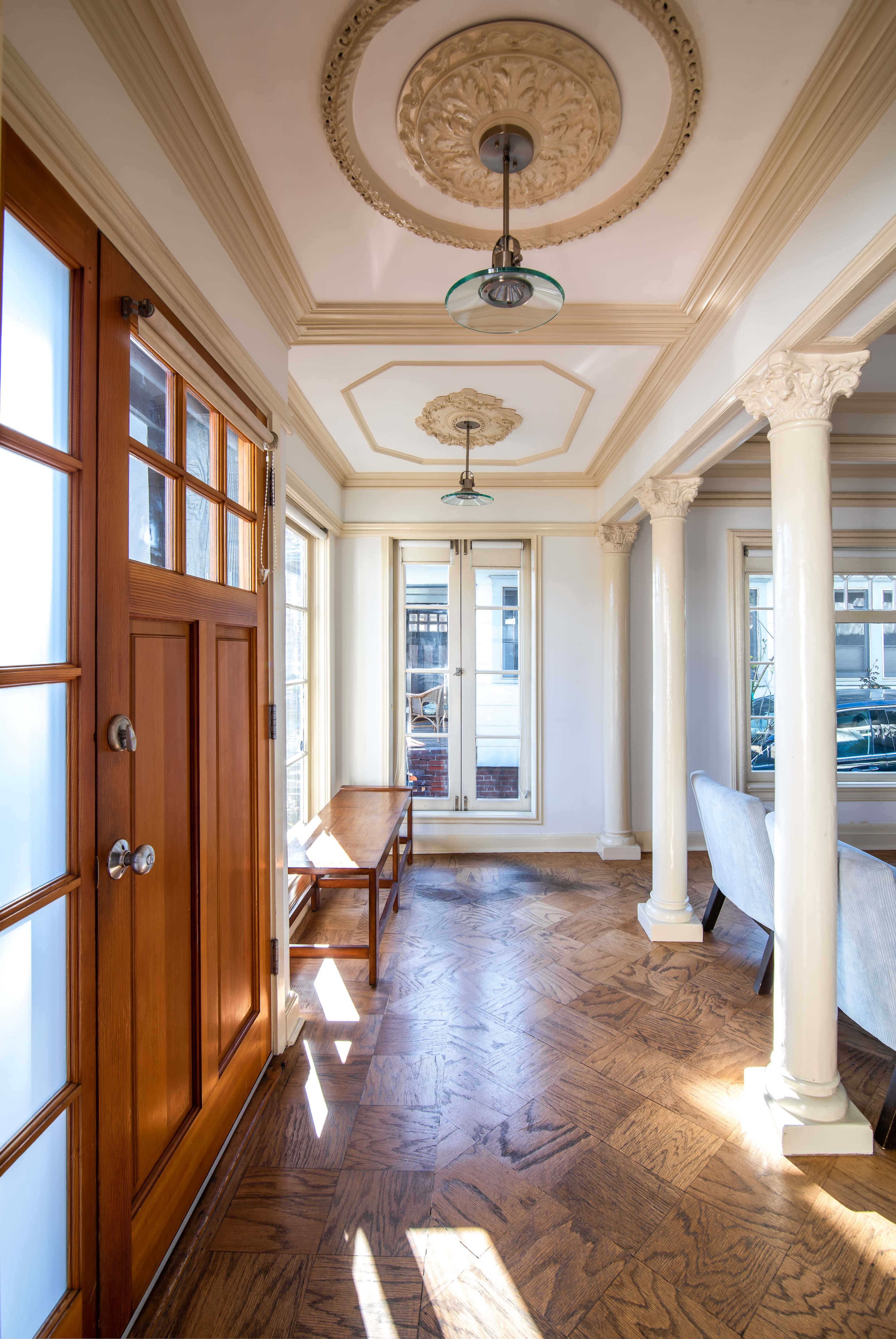 The interior of a well-lit hallway features a wooden front door, ornamental ceiling, and white columns, with a bench and large windows lining the walls.