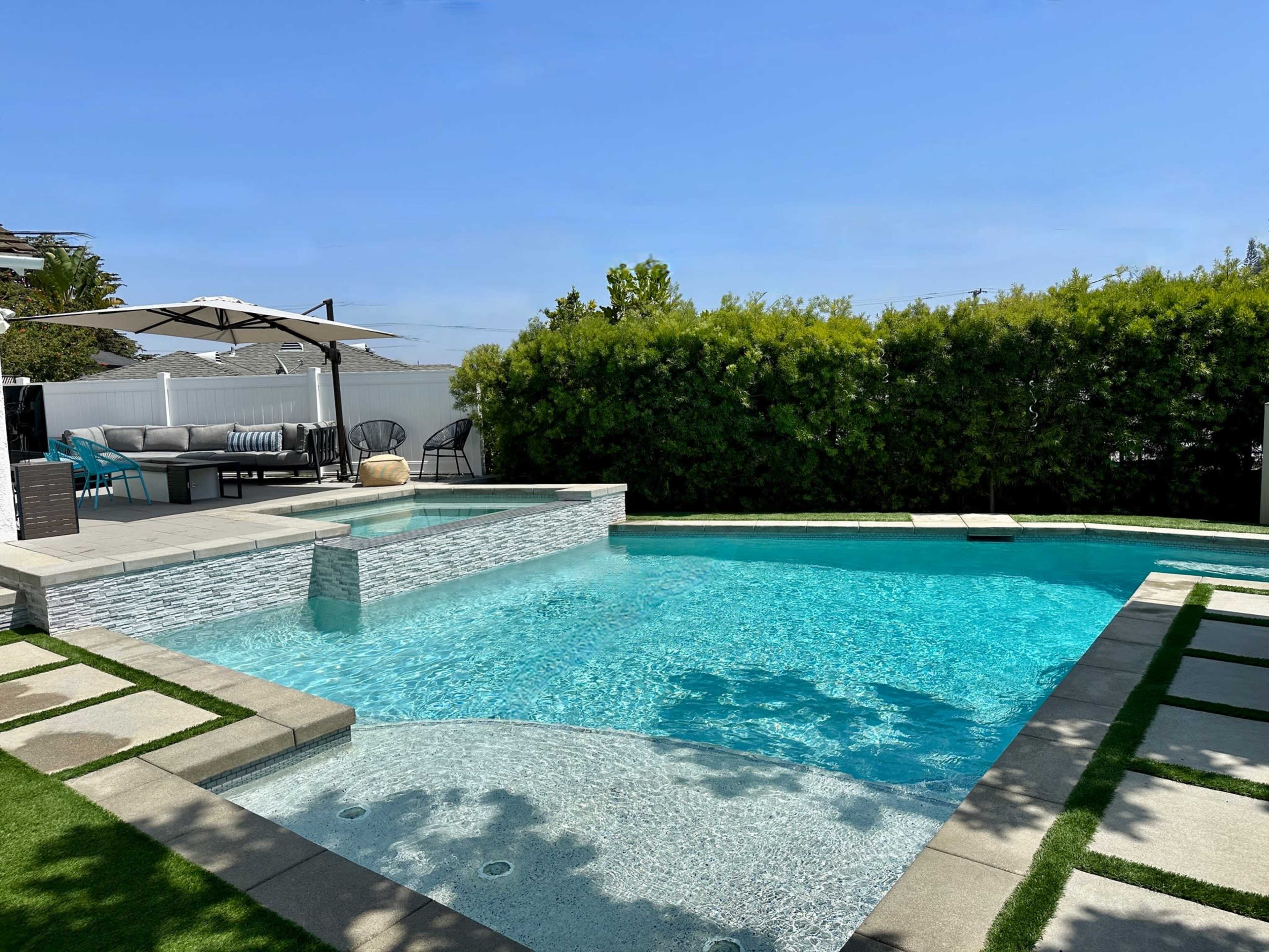 A swimming pool features a stone deck surrounded by greenery and patio furniture under umbrellas.