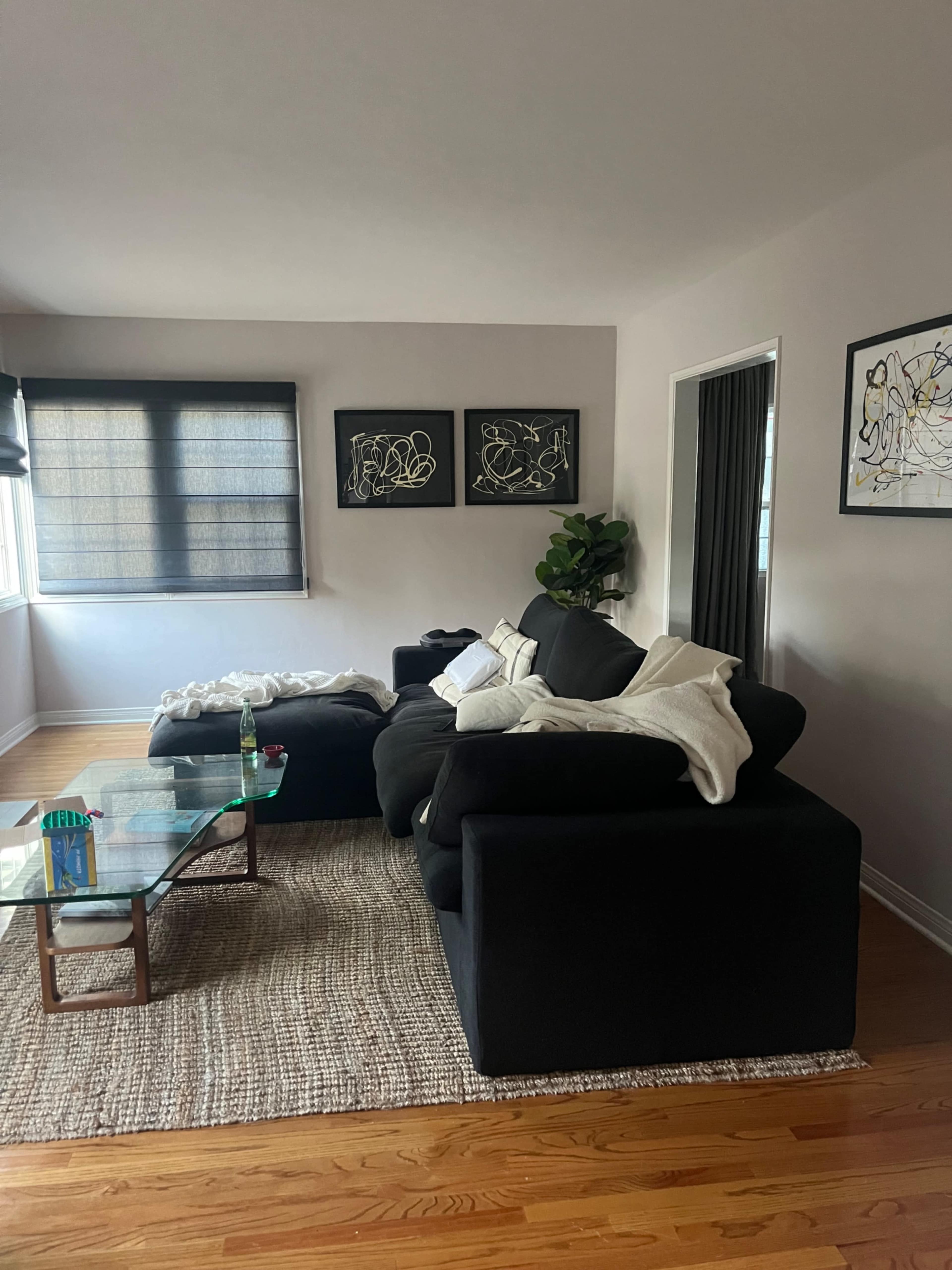 The image shows a modern living room with a black sectional sofa, a glass coffee table, and two framed artworks on the walls.