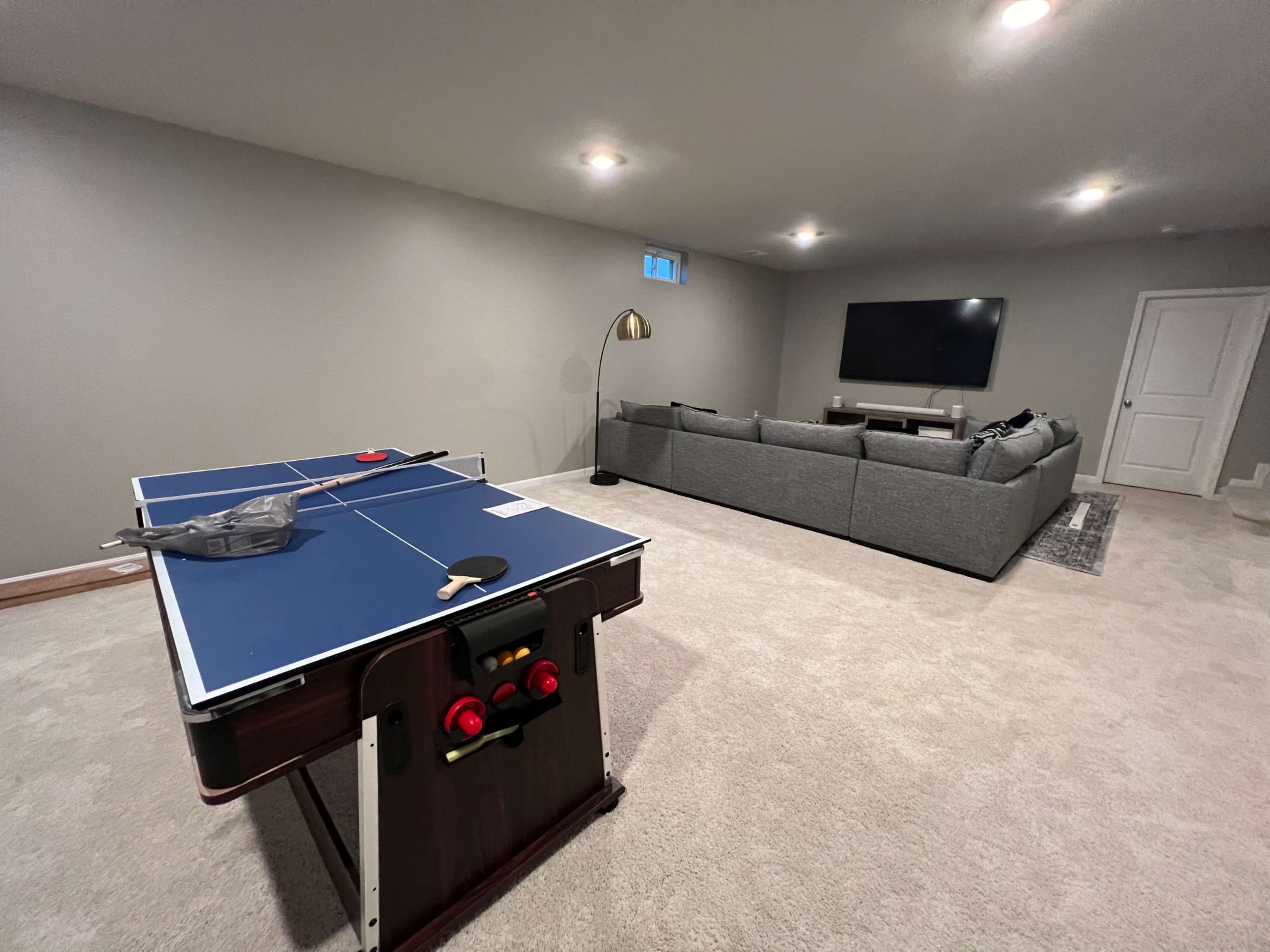 A basement recreation room features a blue ping pong table in the foreground and a gray sectional sofa facing a large wall-mounted TV.