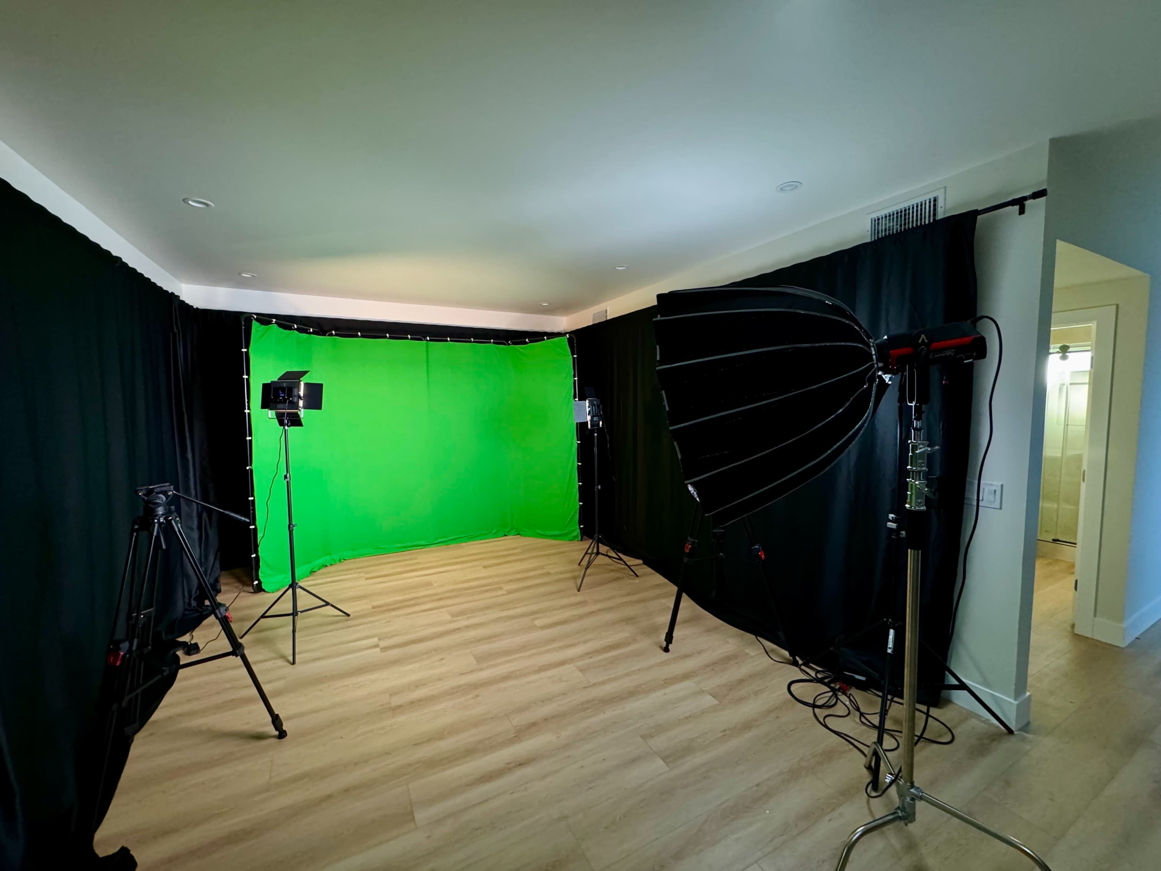 The image shows a room with a green screen setup, black drapery, and lighting equipment arranged for filming.