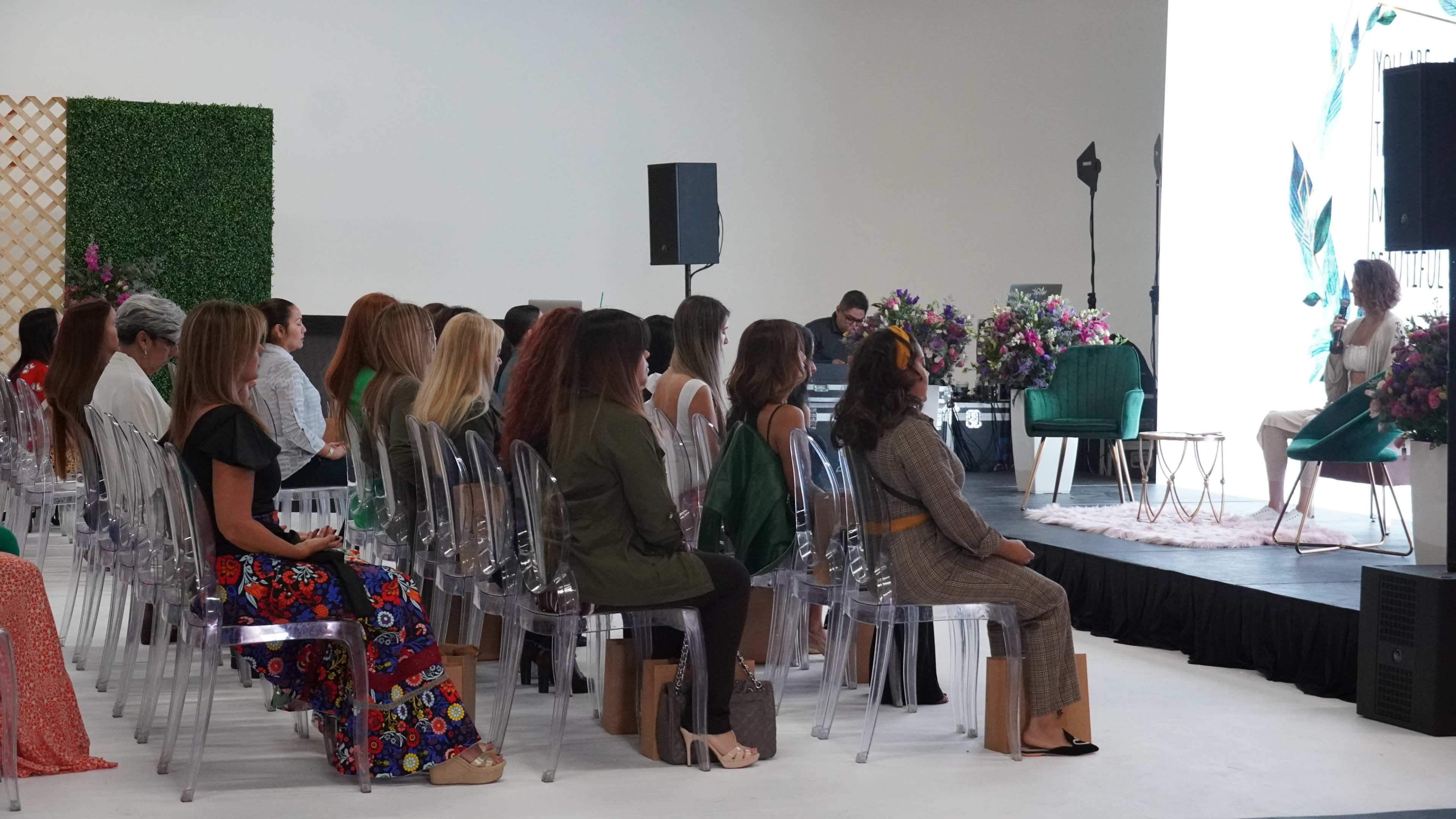 A group of women is seated in clear chairs, attentively watching a presentation on stage at a large indoor event.