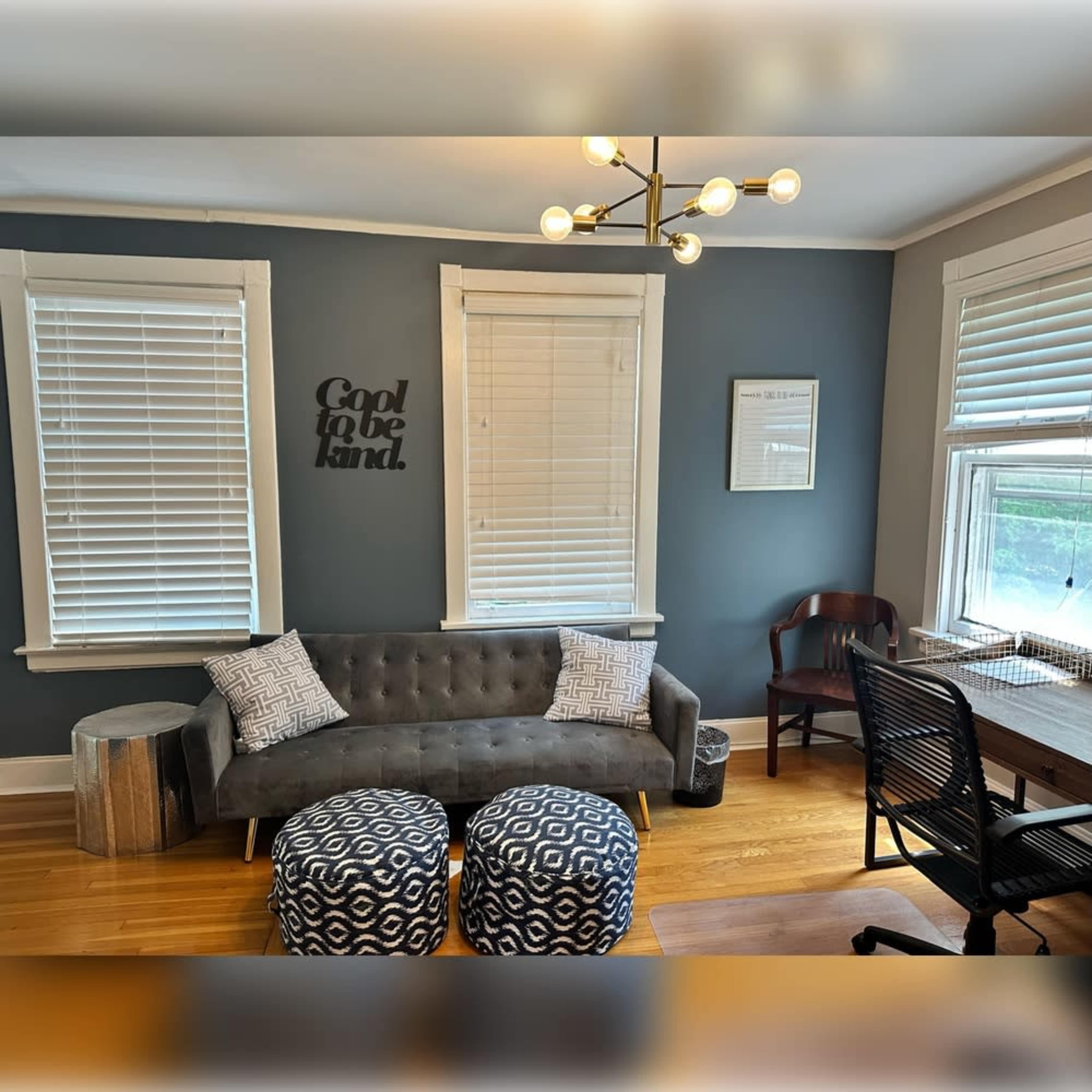 The image shows a well-lit sitting area featuring a gray couch with patterned cushions, two circular ottomans, and a desk with a chair against a blue wall.