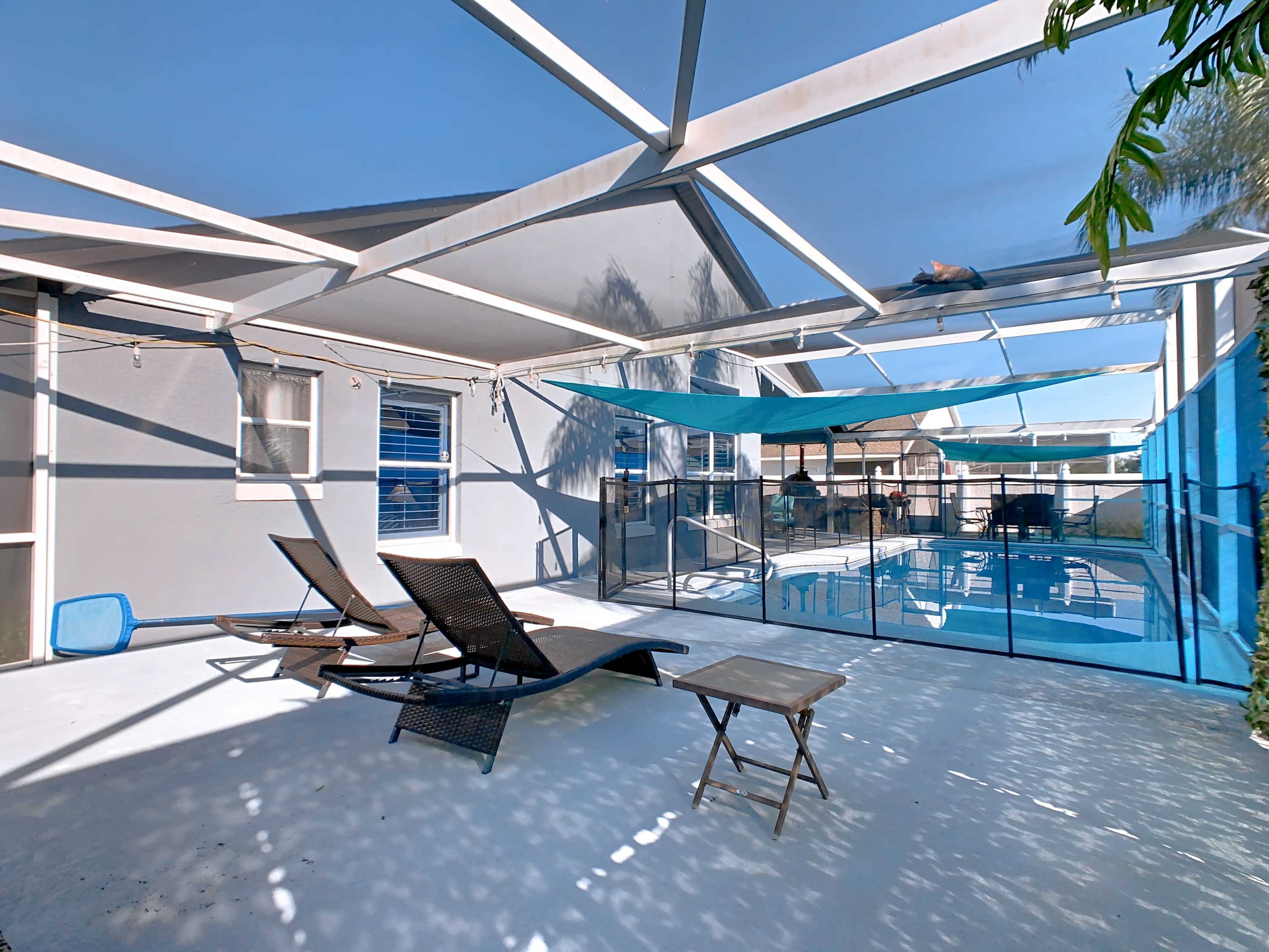 The image shows a patio area with lounge chairs, a small table, and a pool enclosed by a safety fence, all under a blue sky and a covered pergola structure.
