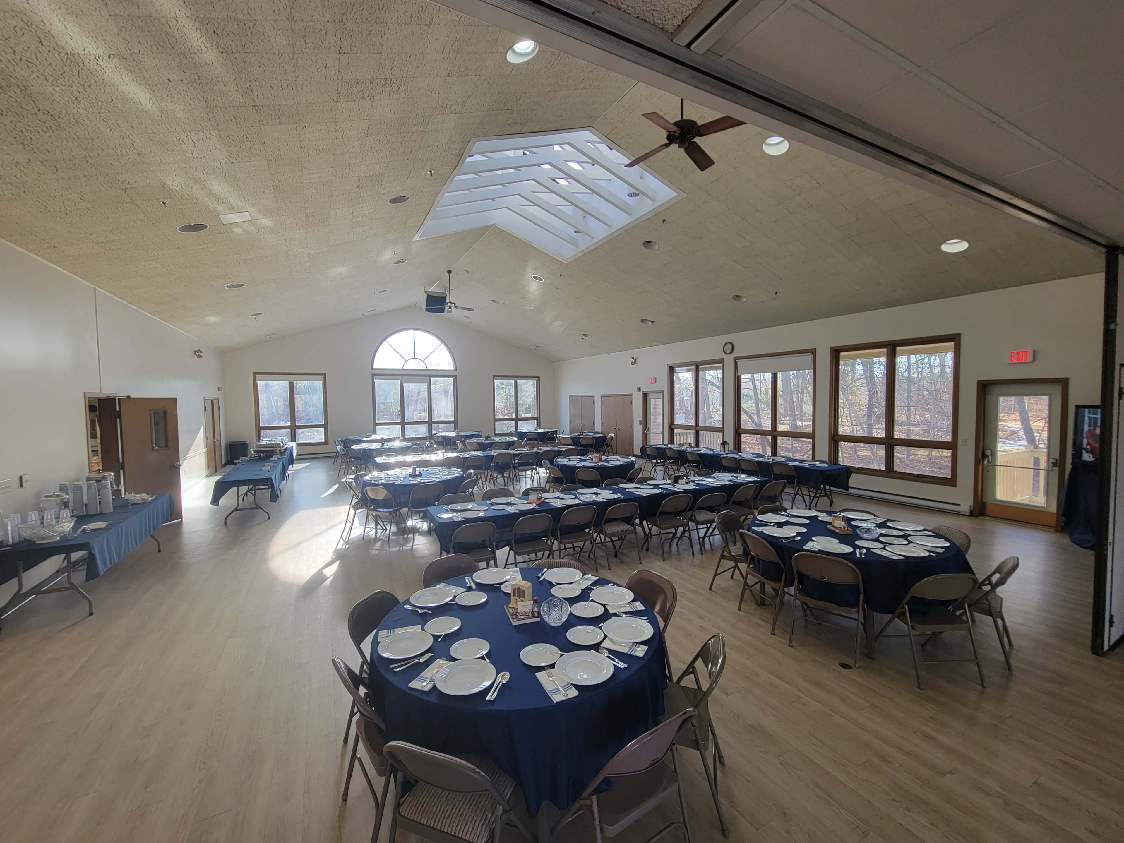 The image shows a spacious banquet hall arranged with several round tables set with blue tablecloths and dinnerware, featuring large windows that allow natural light to enter.