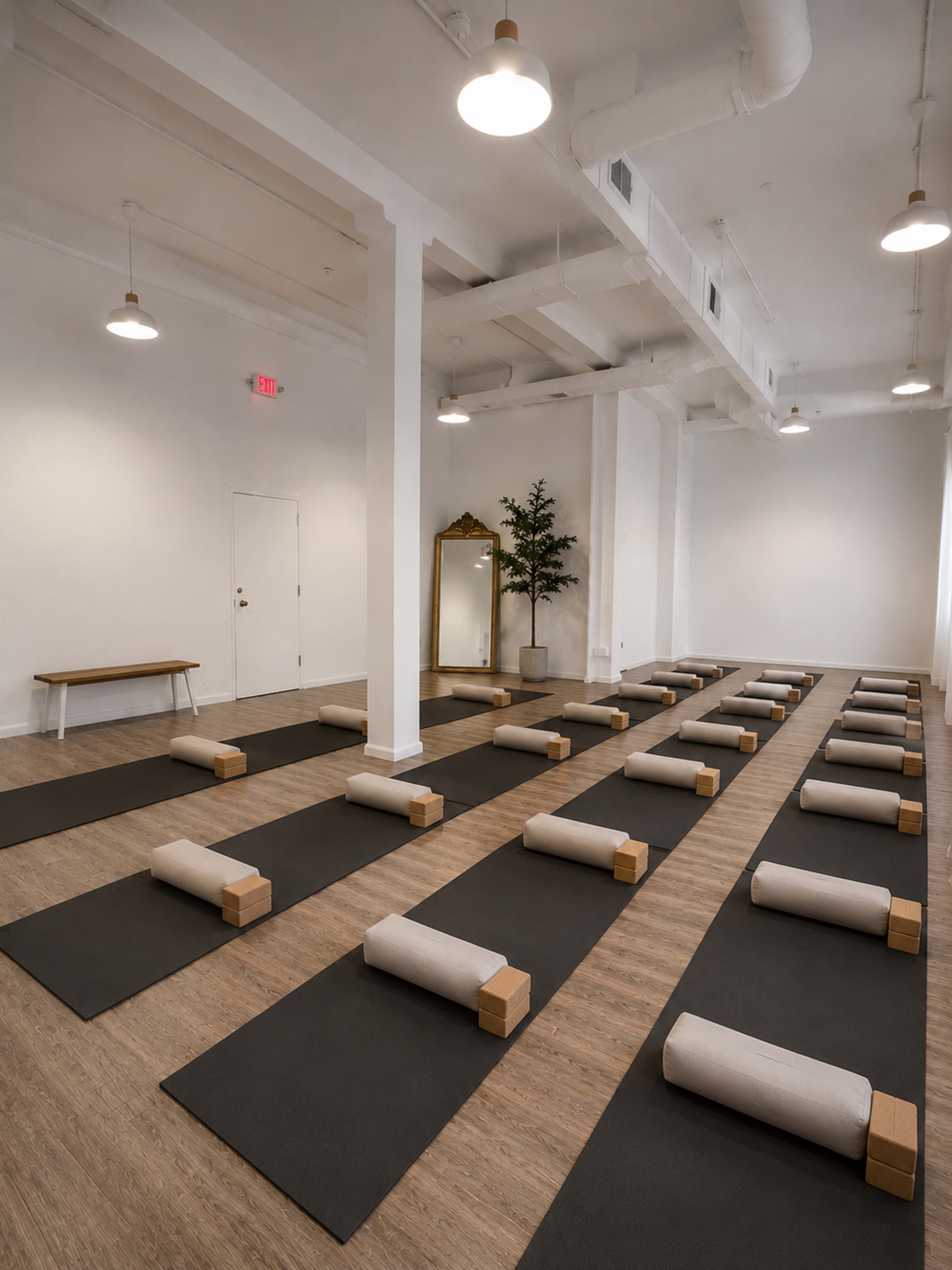 A spacious yoga studio features neatly arranged mats with wooden blocks and mirrors, illuminated by soft overhead lighting.