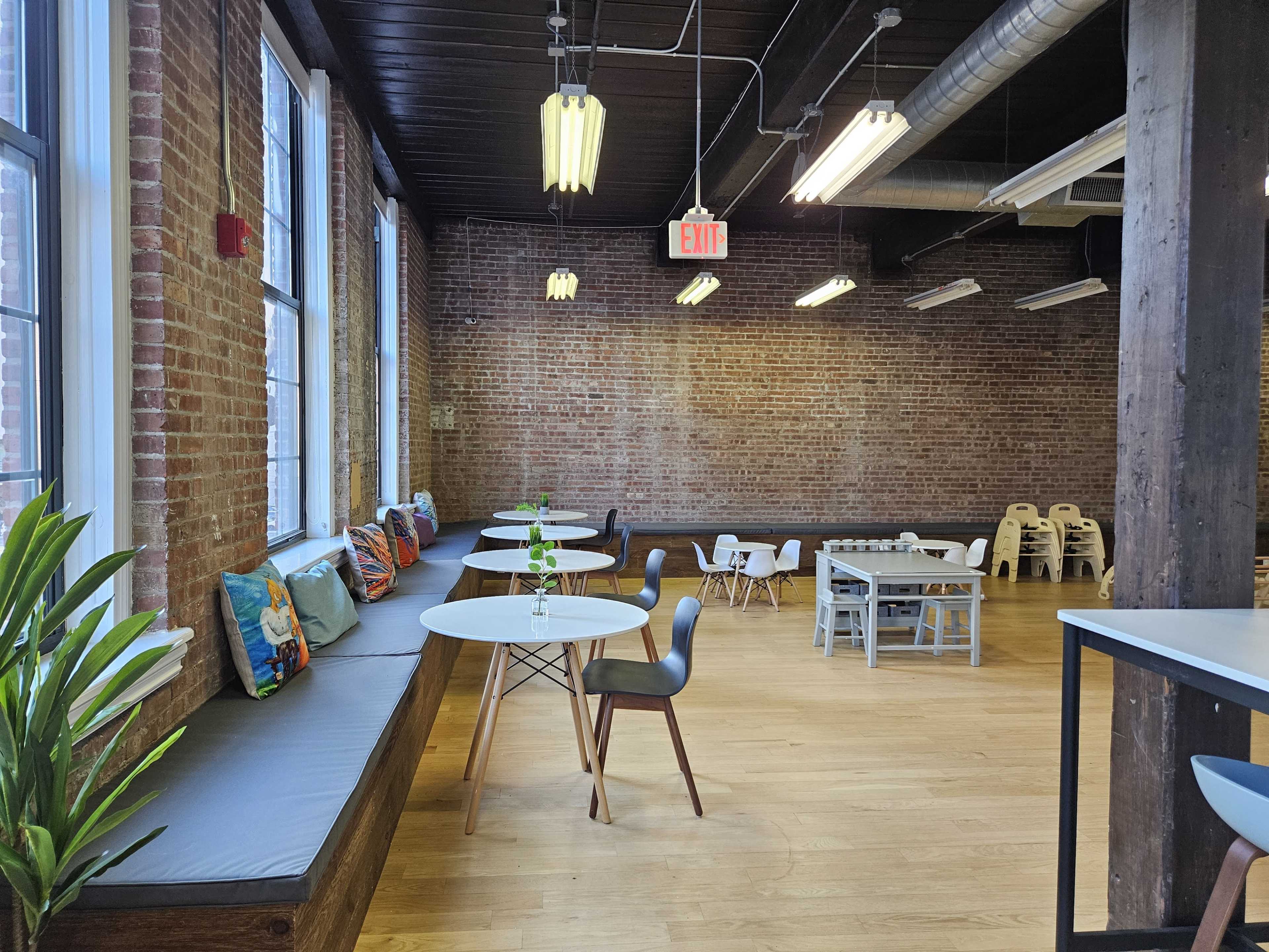 The image shows a spacious room with brick walls, wooden floors, and a mix of seating arrangements, including tables and chairs, as well as a window bench with cushions.