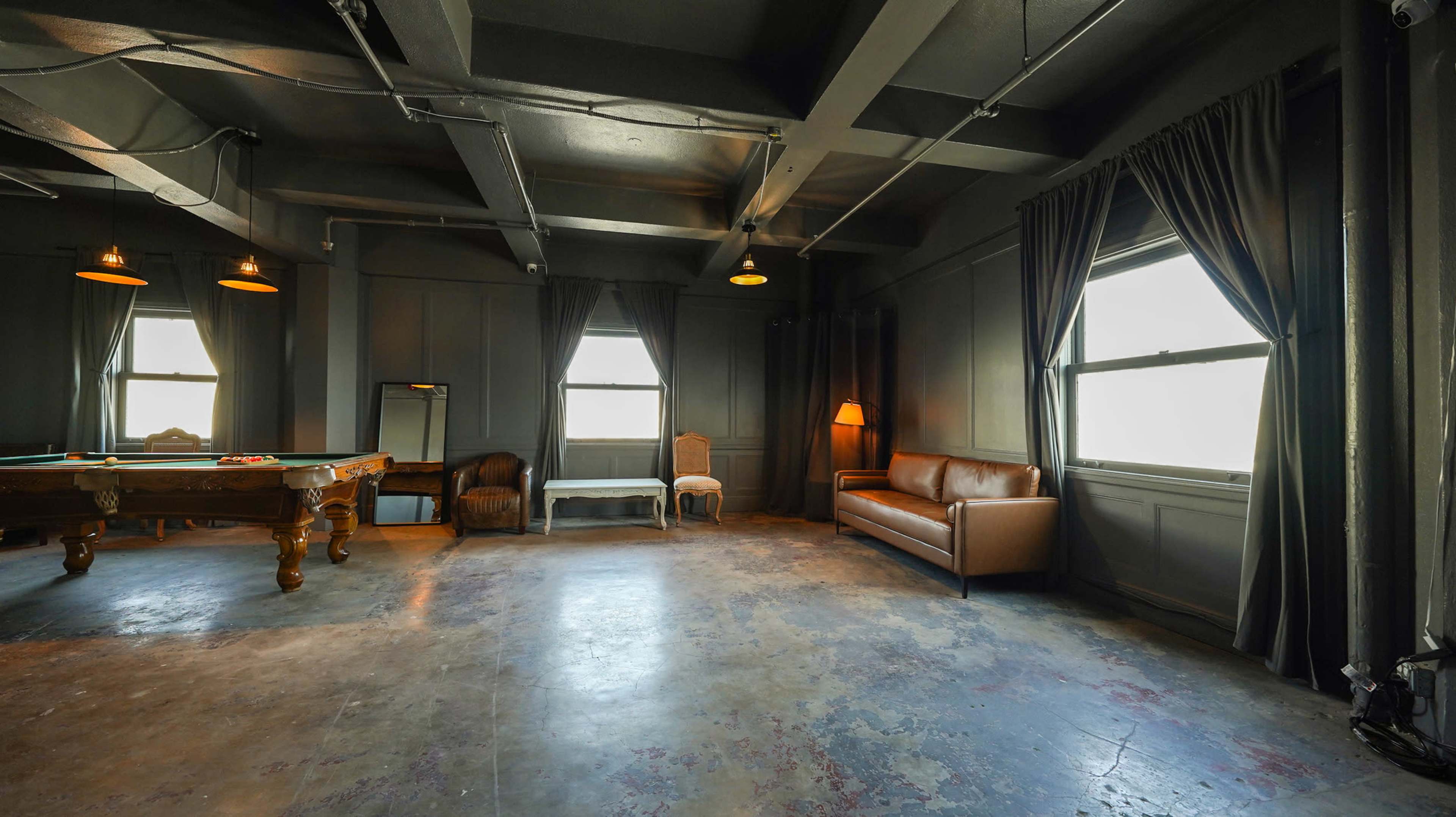 A dimly lit room with a pool table, a brown leather sofa, and several windows covered with dark curtains.
