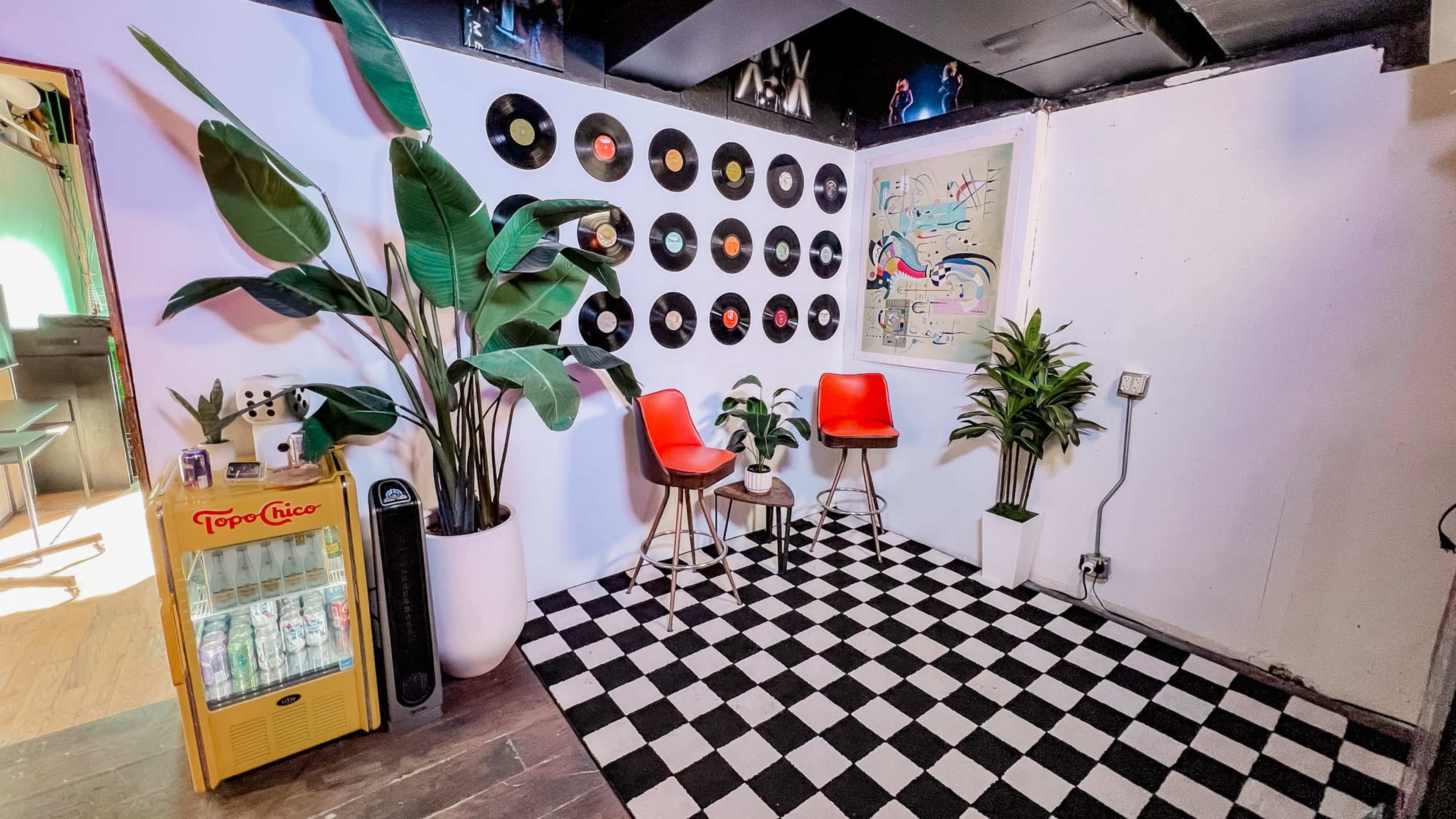 The scene features a stylish indoor space with a black and white checkered floor, two red chairs on wooden stools, a large potted plant, and a wall decorated with vinyl records and art.