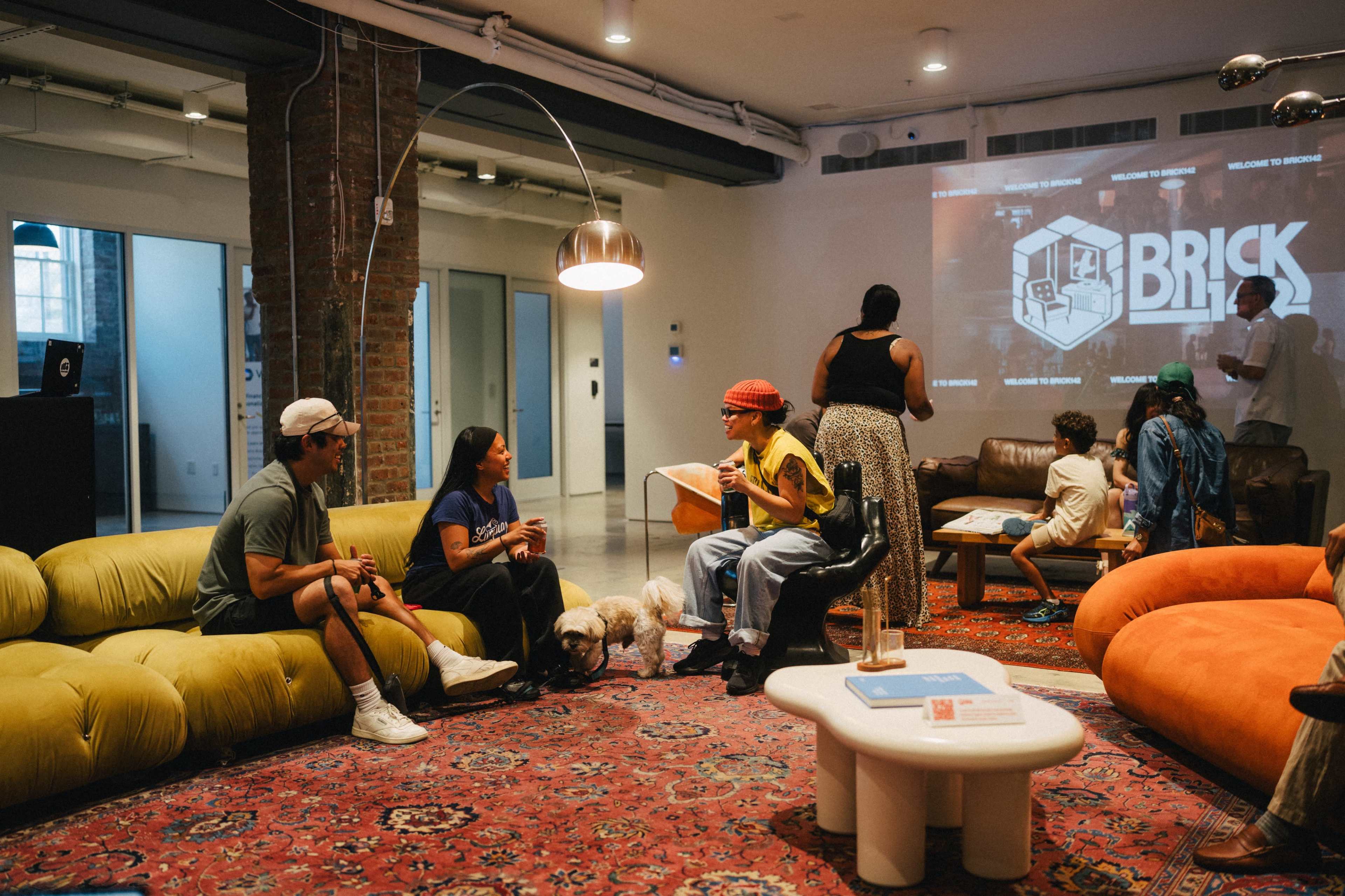 A group of people is engaged in conversation in a cozy, modern lounge area featuring colorful furniture and a large rug.