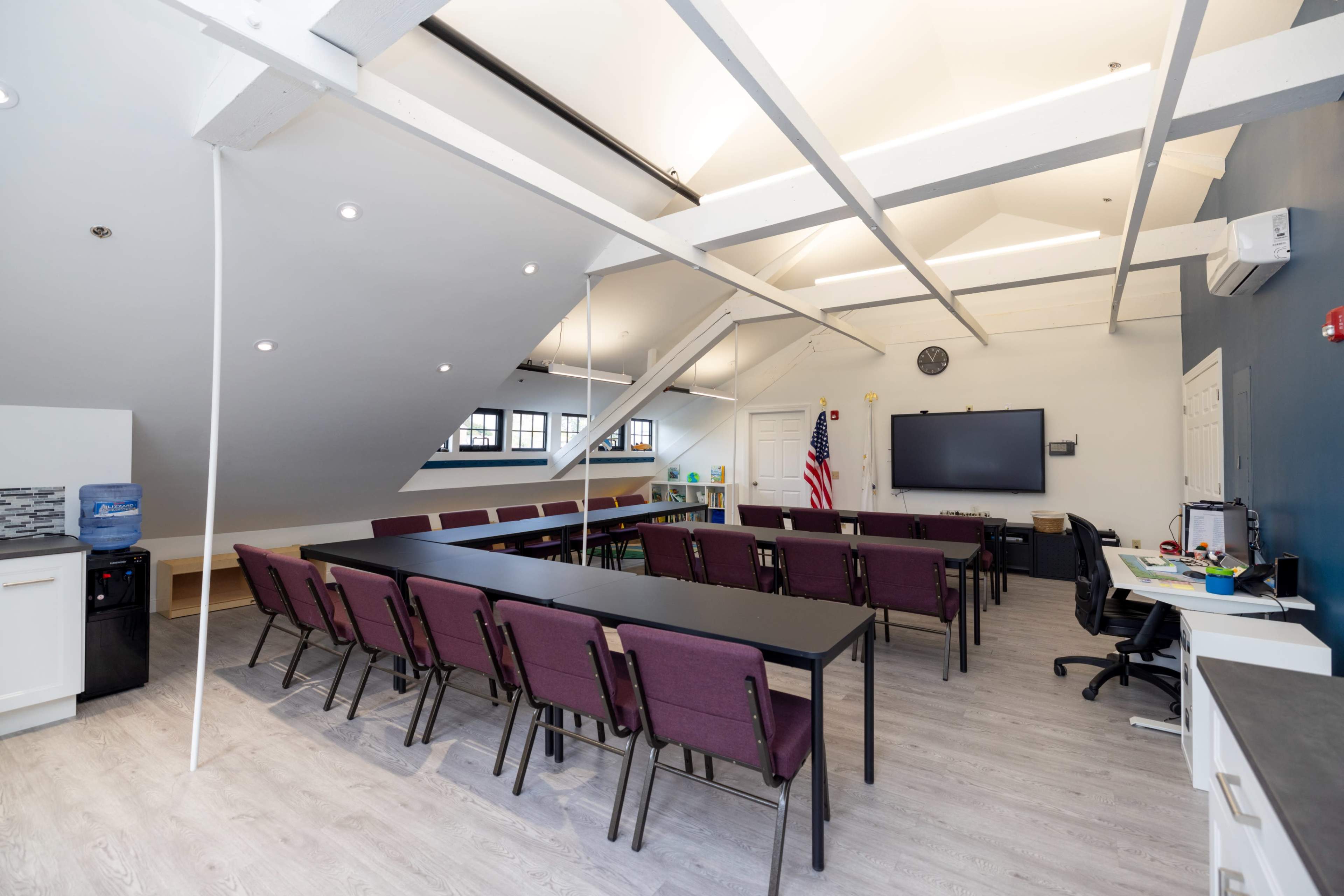 A bright conference room with a long arrangement of tables and chairs, a large screen on the wall, and an American flag in the corner.