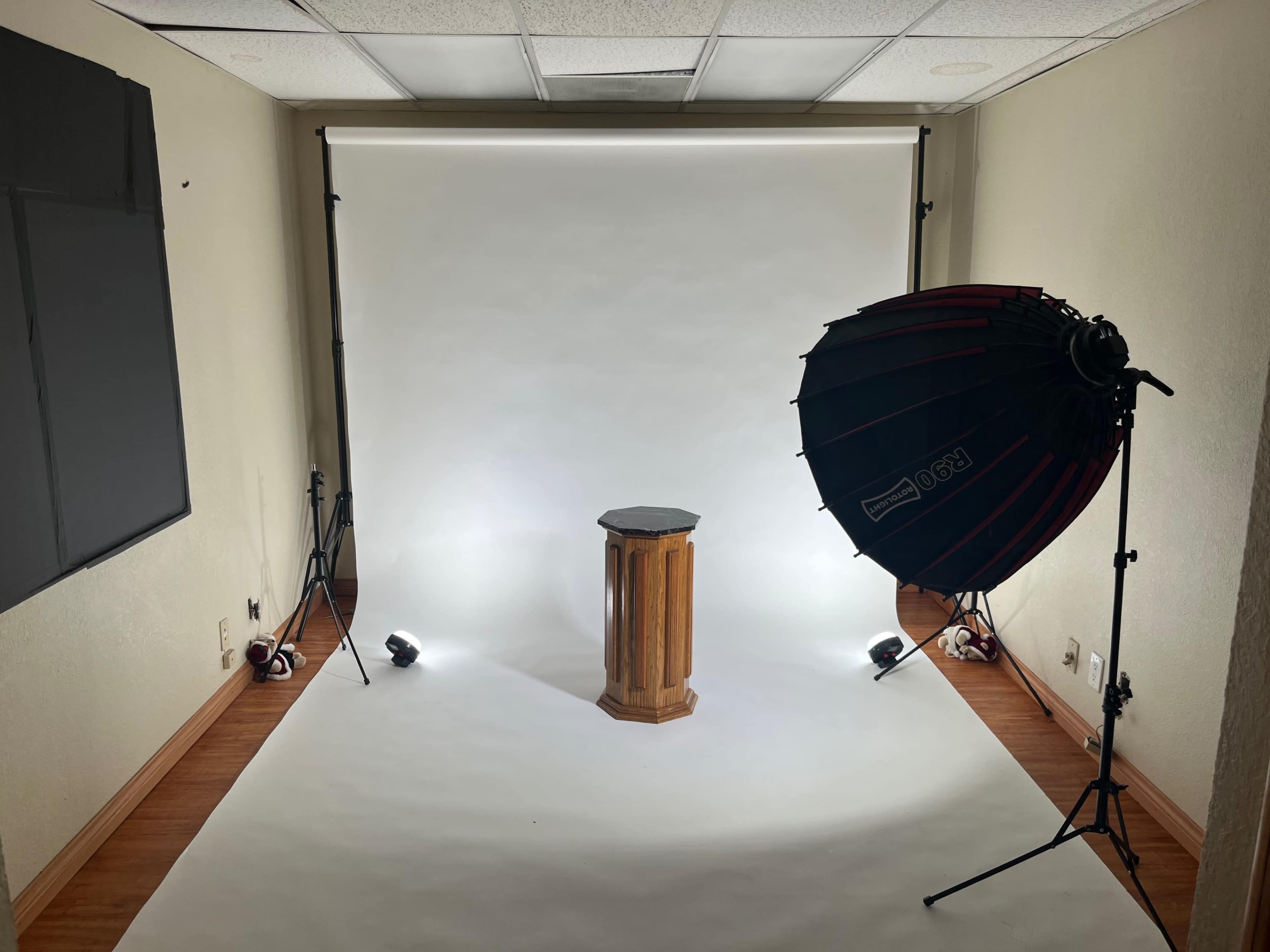A photography studio with a wooden pedestal in the center, surrounded by softbox lighting and a plain white backdrop.