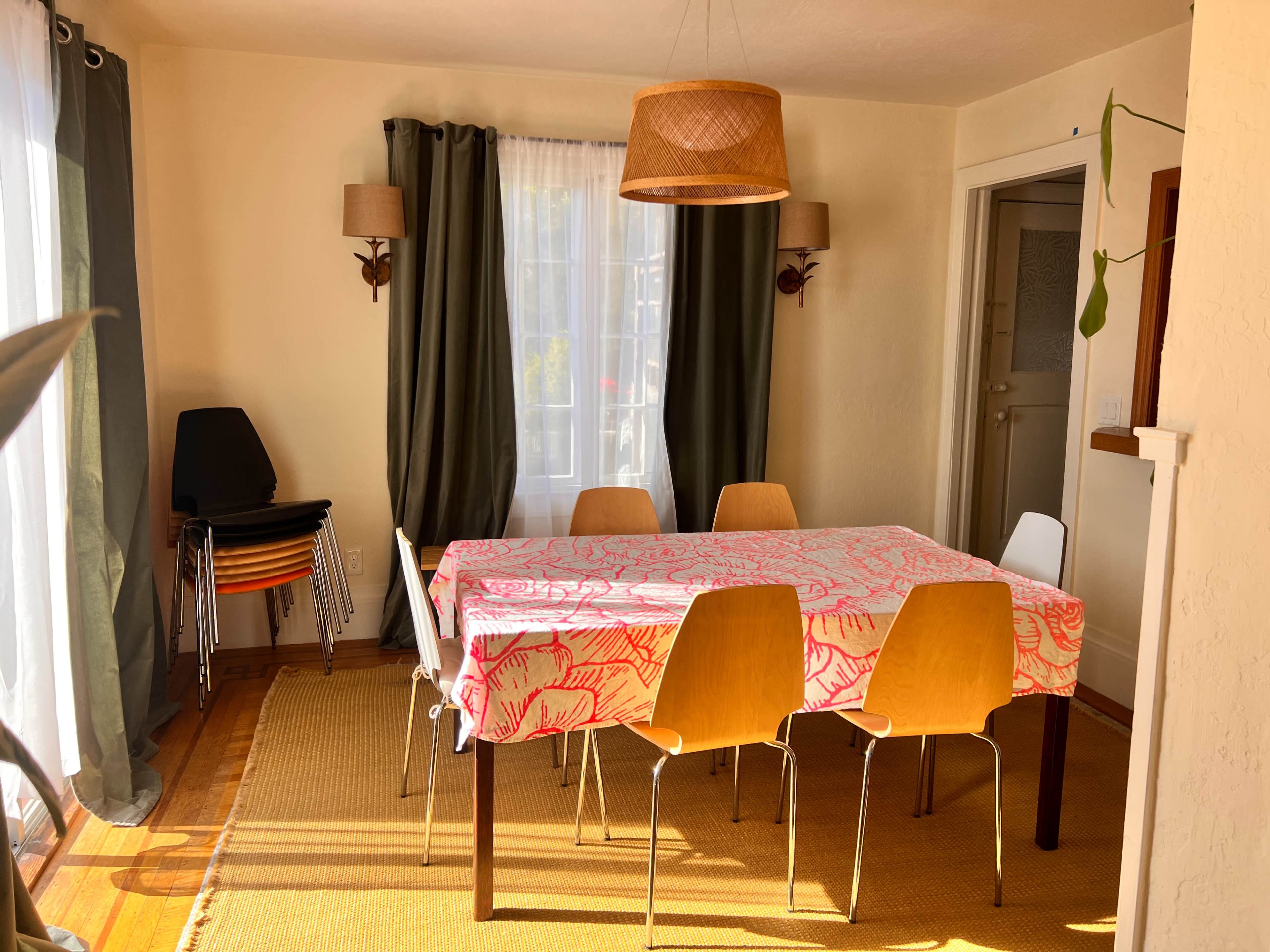 The image shows a dining area with a table covered by a patterned tablecloth, surrounded by six chairs, a woven light fixture hanging above, and large windows with curtains allowing natural light to enter.