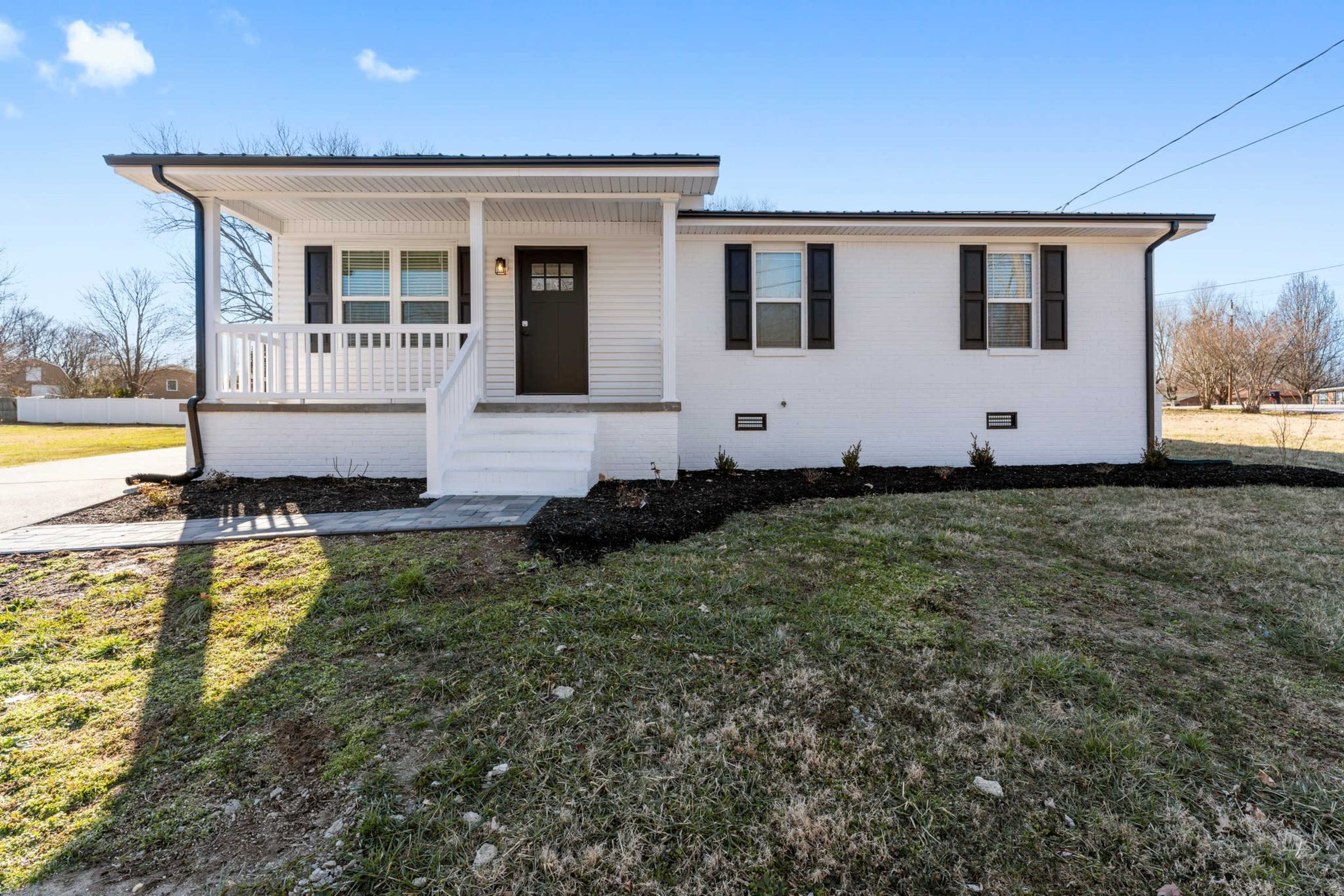 A single-story, white brick house with a front porch, black shutters, and a landscaped yard.