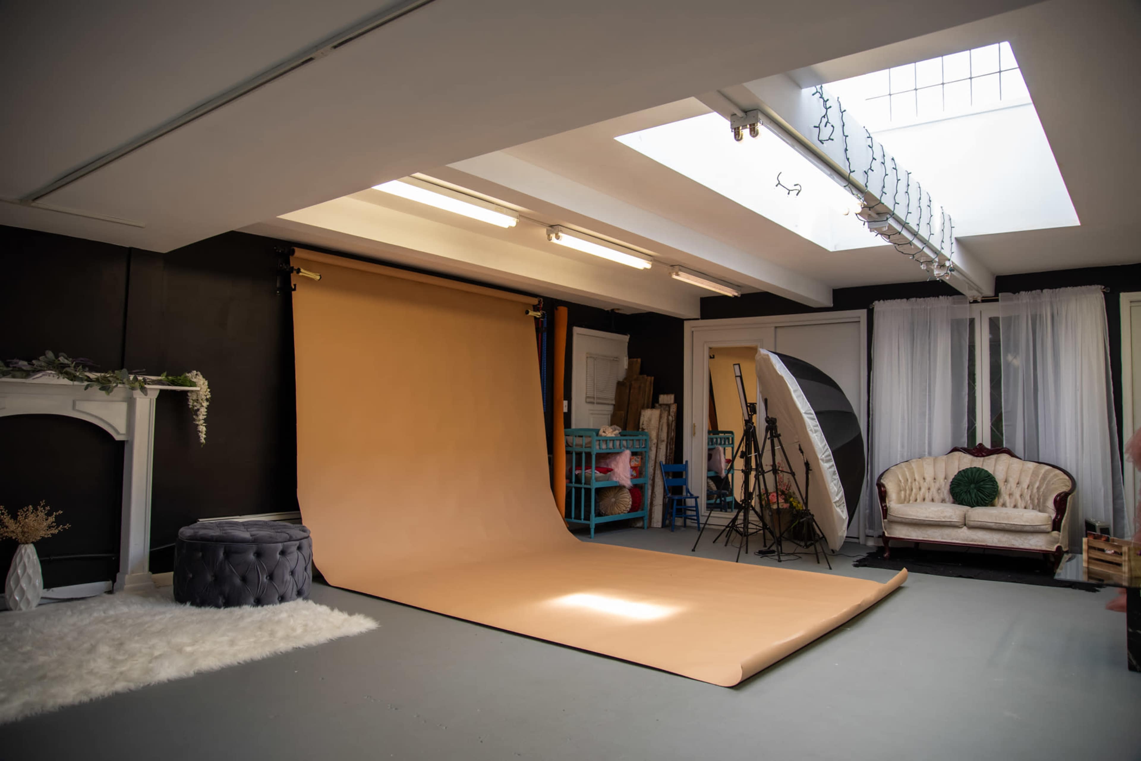 A photography studio featuring a large beige backdrop, soft lighting equipment, and vintage furniture in a well-lit room.