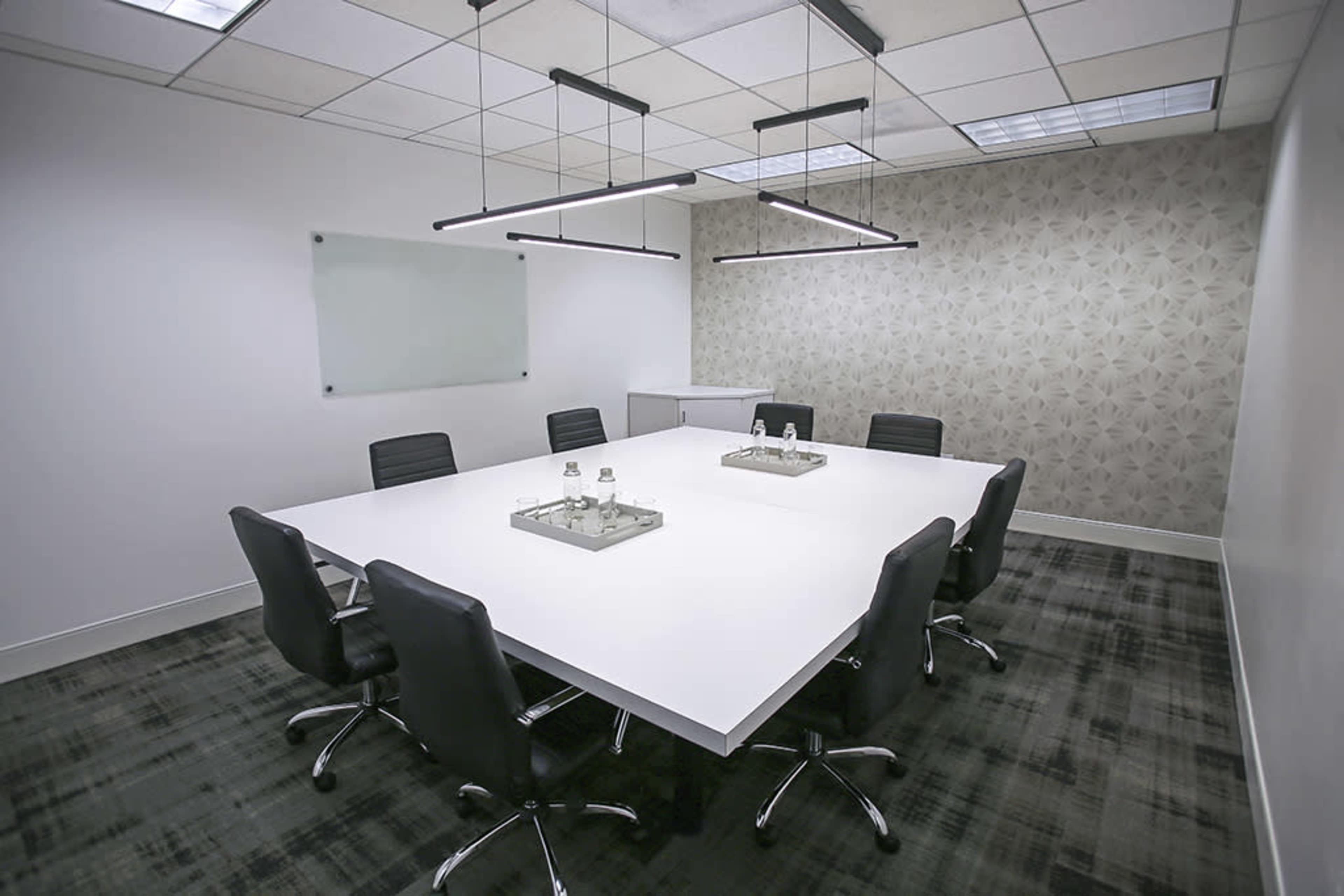 A modern conference room features a large white table surrounded by black chairs, with minimalistic light fixtures above and a patterned wall in the background.