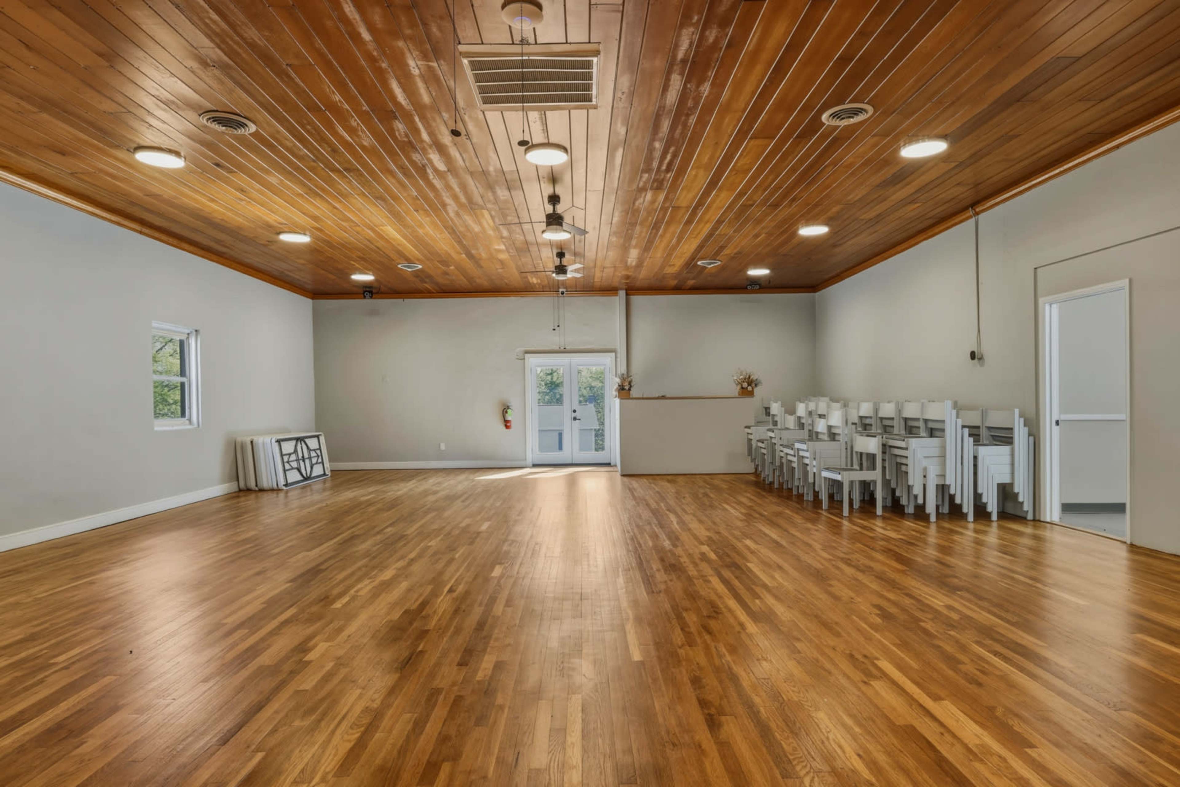 The image shows a spacious room with wooden flooring and a ceiling adorned with wooden panels, featuring a row of white chairs stacked against one wall.