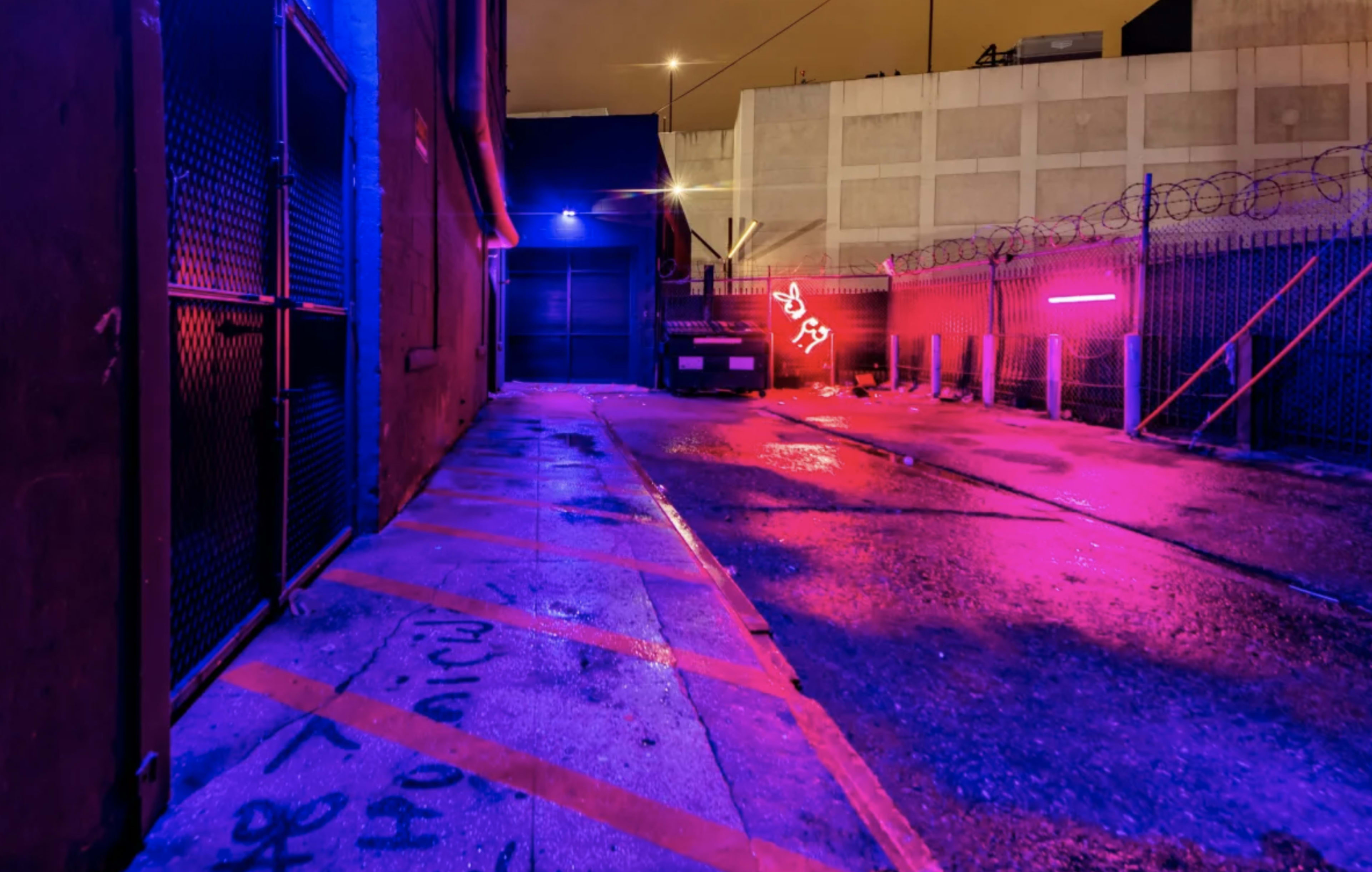 The scene depicts a wet alleyway illuminated by purple and blue neon lights, with a parked vehicle and a barbed-wire fence in the background.
