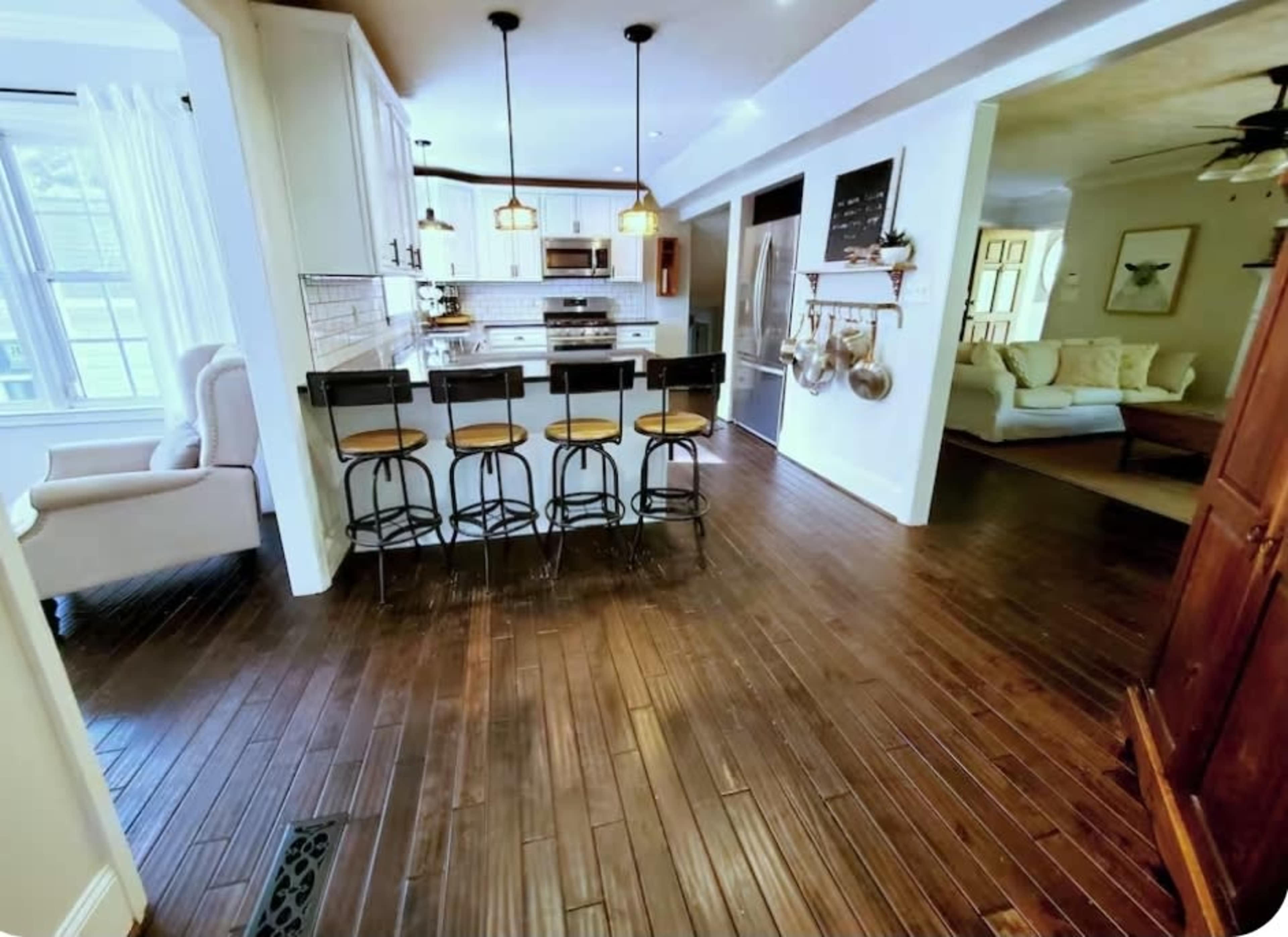 The image shows a modern kitchen with wooden flooring, bar stools at the counter, and an adjacent living area visible through an open doorway.